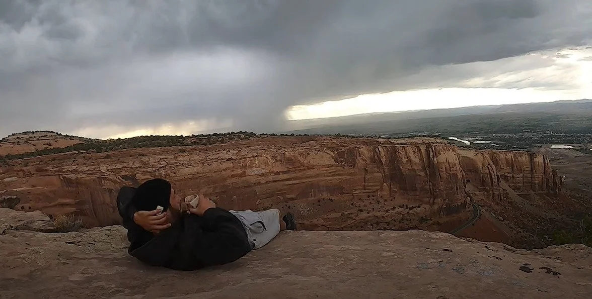 Person lying on a rock ledge, drinking from a cup, overlooking a canyon with dark clouds overhead.