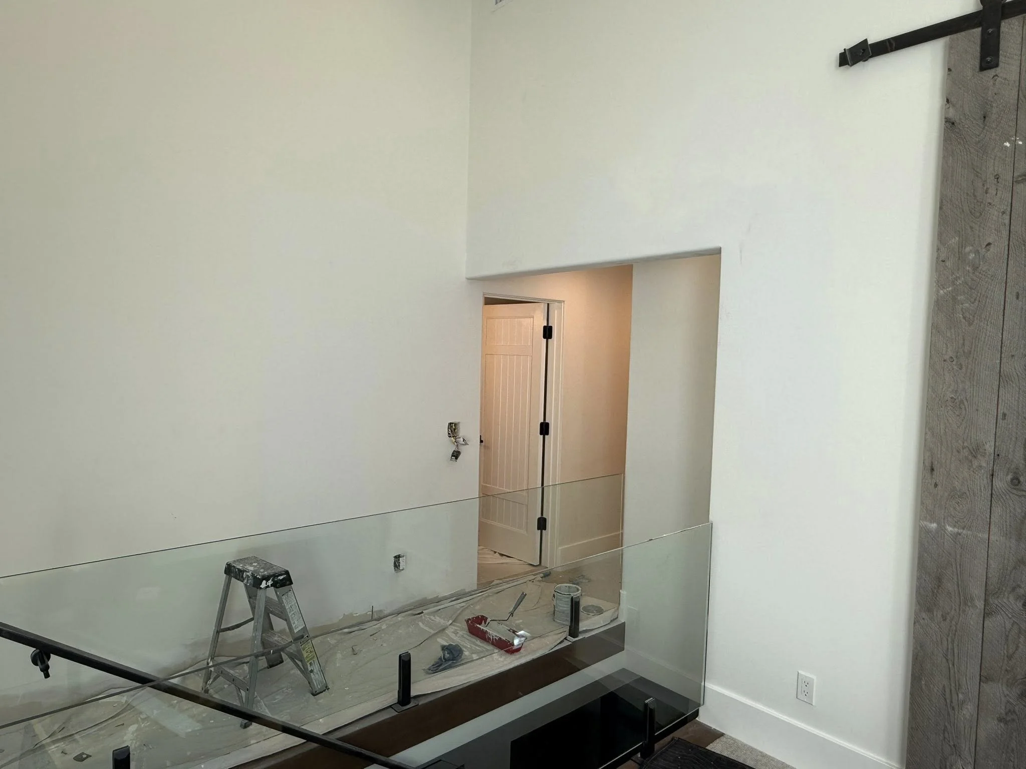 Interior of a room under construction or renovation with a glass railing, a ladder, and tools, leading to a door in the background.