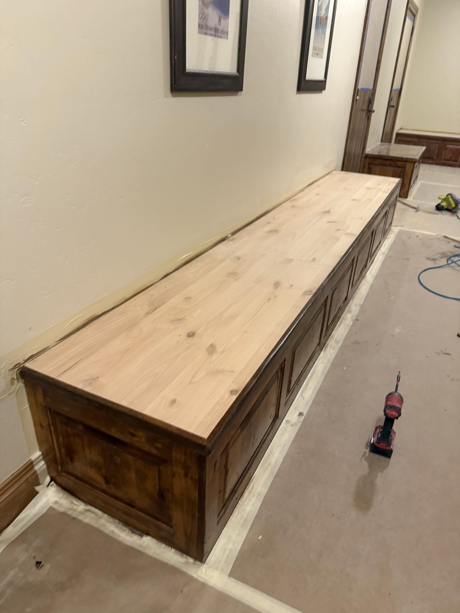 A long wooden cabinet with a fresh natural wood top is being installed along a beige wall in a hallway, with framed artwork above and a door in the background. Tools and construction materials are on the floor nearby.