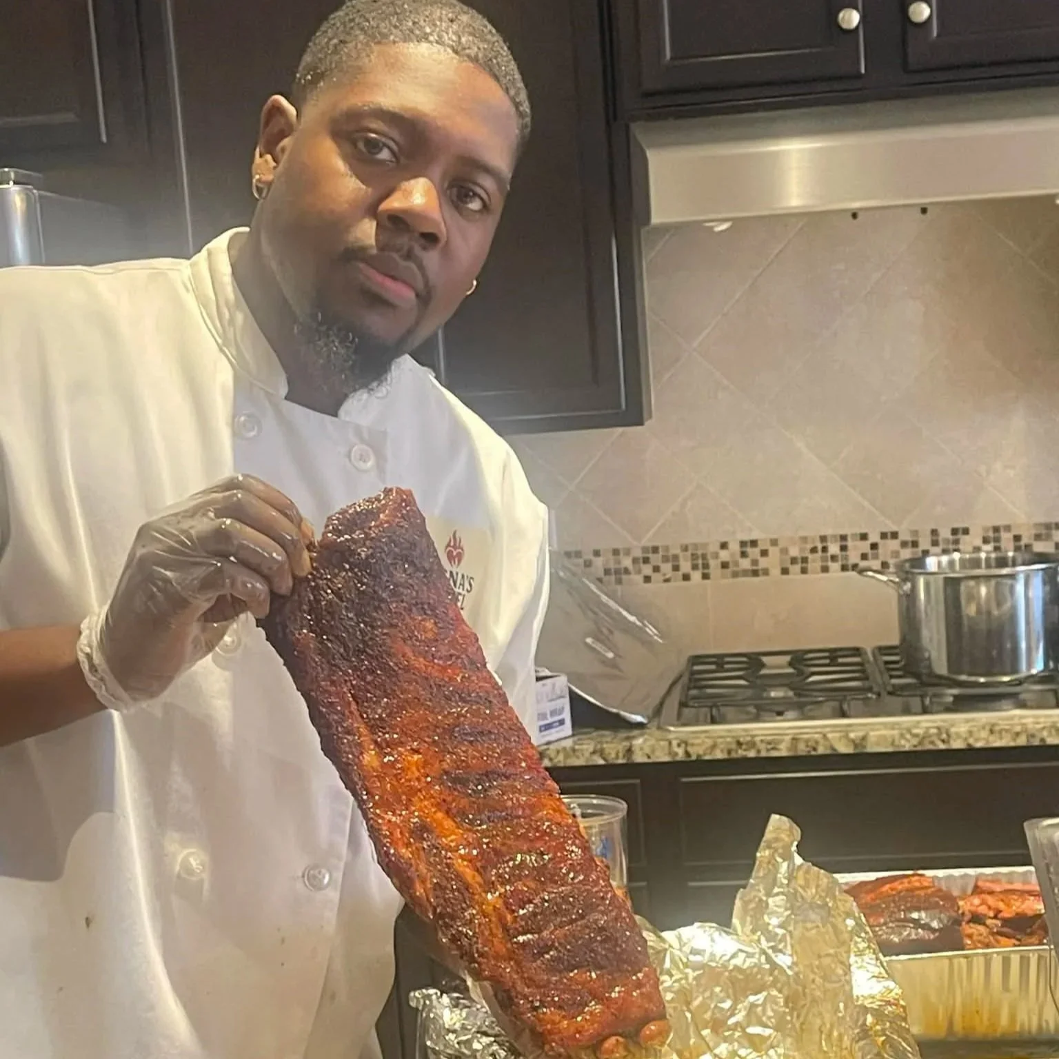 A man in a chef's coat holding a rack of glazed ribs in a kitchen.