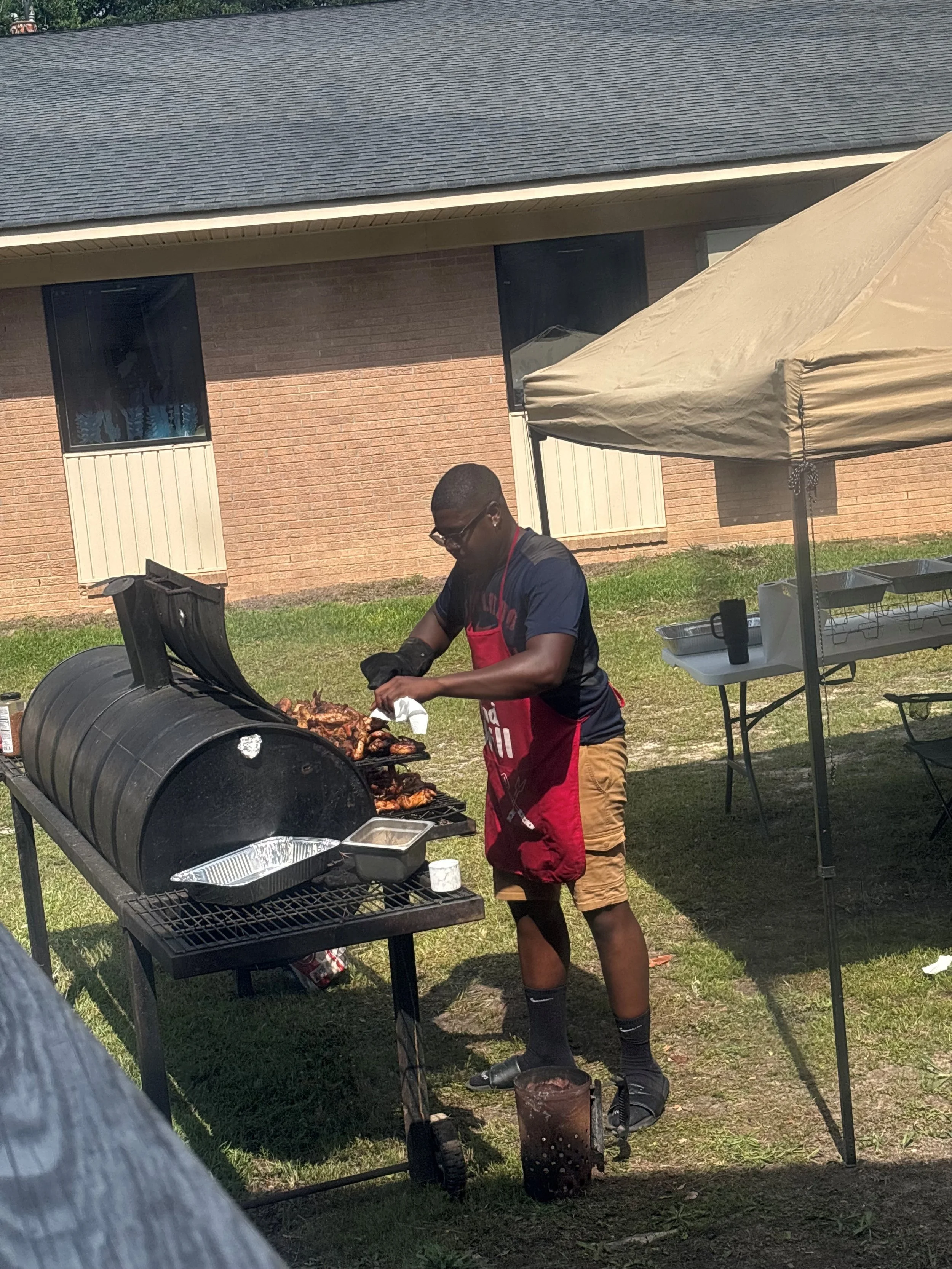 Young man grilling meat outdoors, wearing black gloves, sunglasses, a black and blue shirt, khaki shorts, black socks, and a red apron under a beige canopy tent with tables and trays nearby.