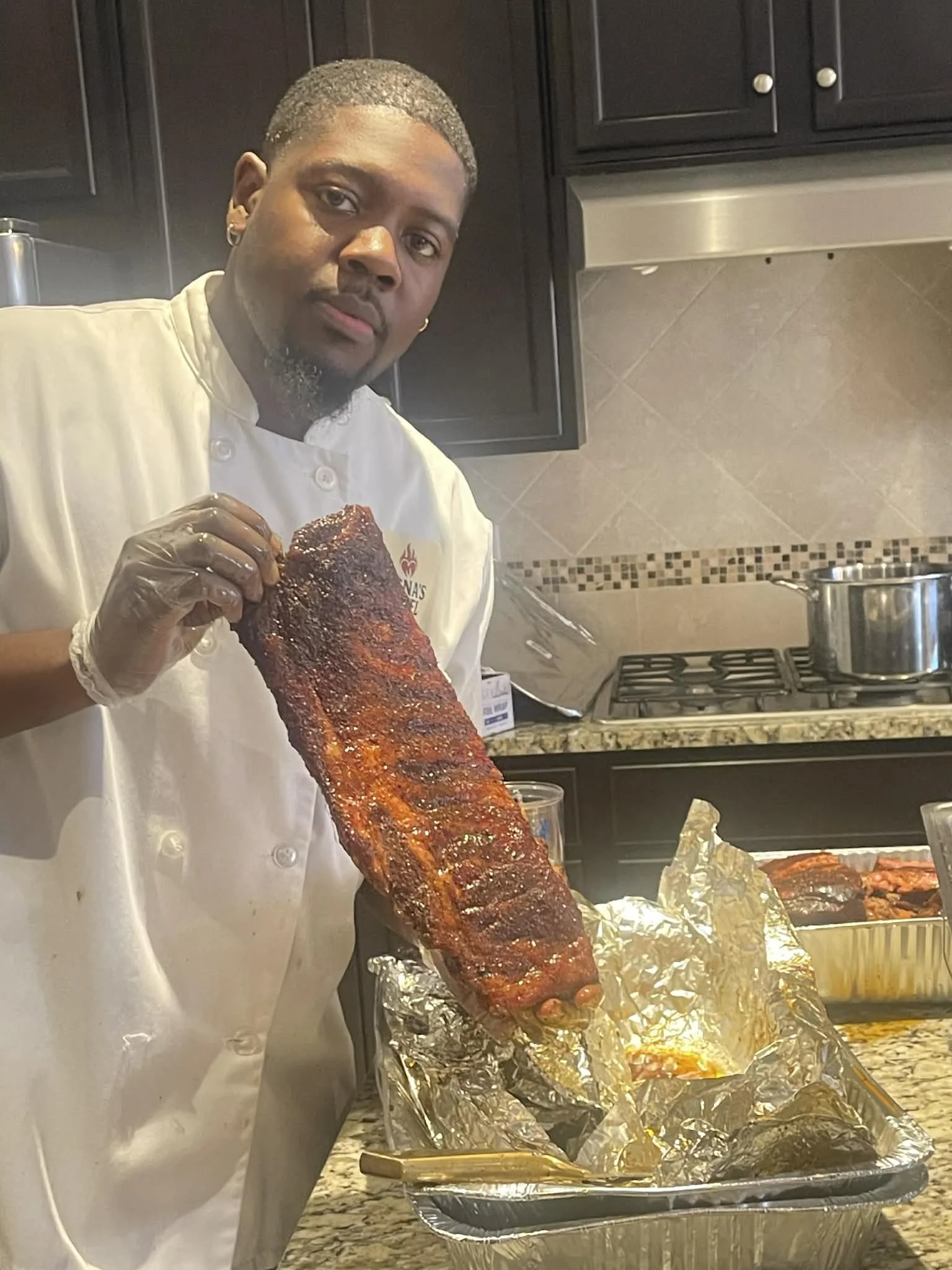A man wearing a chef's coat and gloves holding a rack of smoked or grilled ribs in a kitchen.