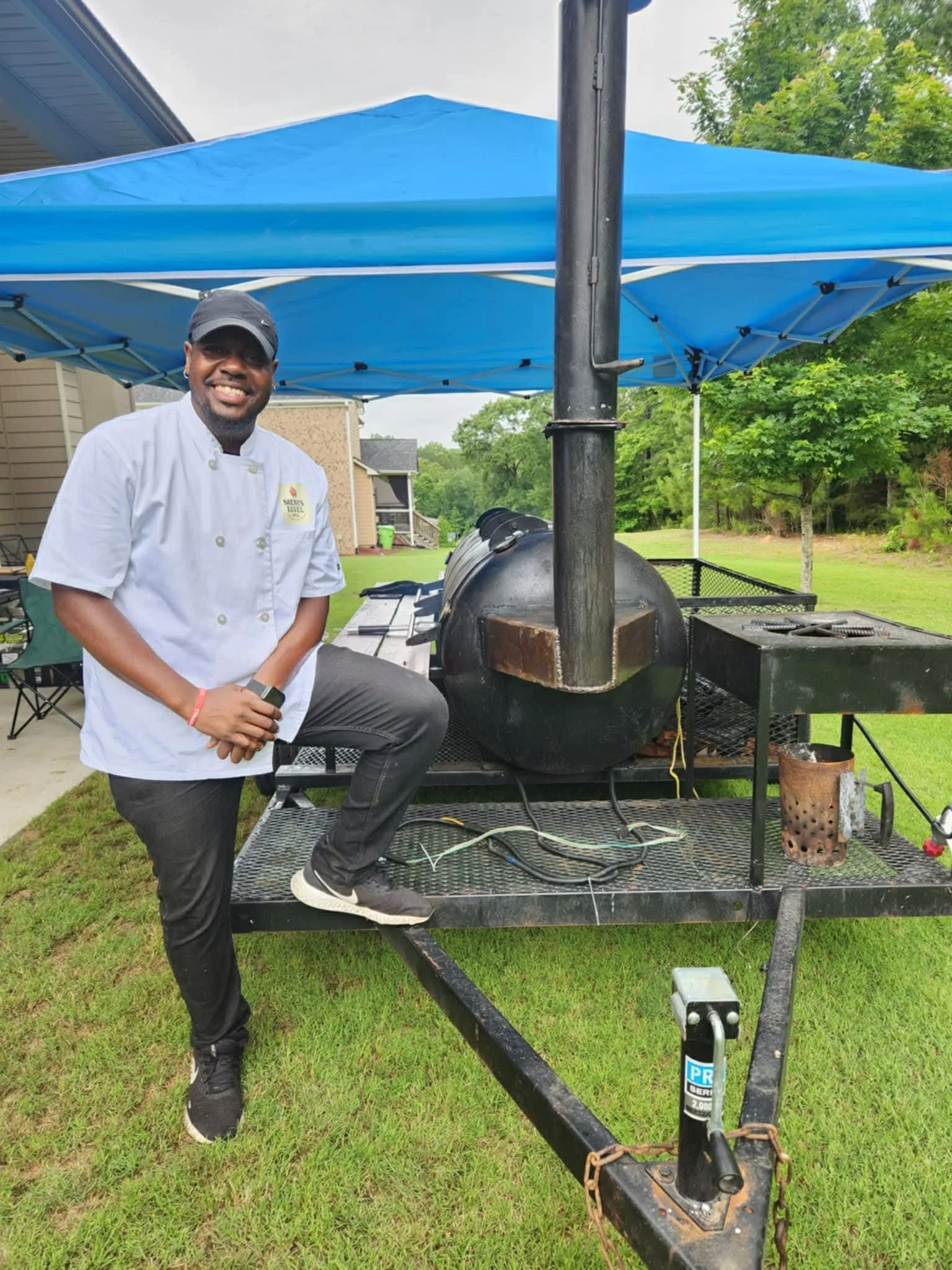 A man in a white chef's coat and black pants poses with one foot on a barbecue smoker on a trailer, under a blue canopy tent in a backyard with green grass and trees.