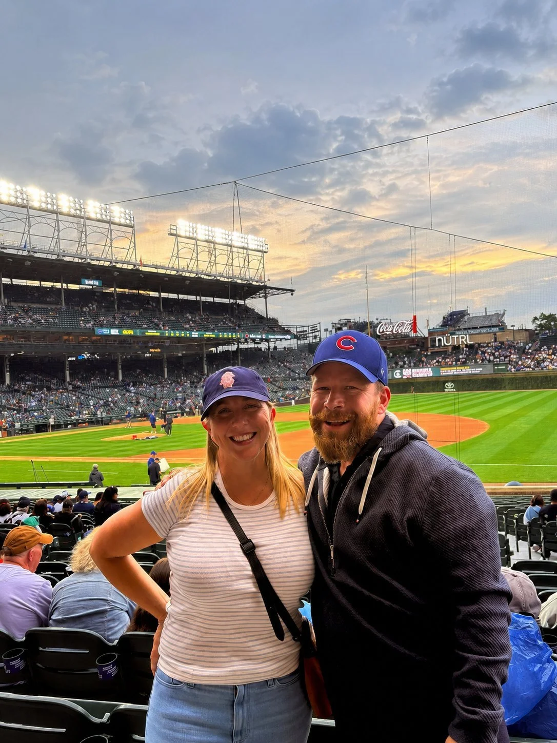 A smiling woman and man at a baseball stadium during sunset, both wearing Chicago Cubs hats, with the field and other fans in the background.