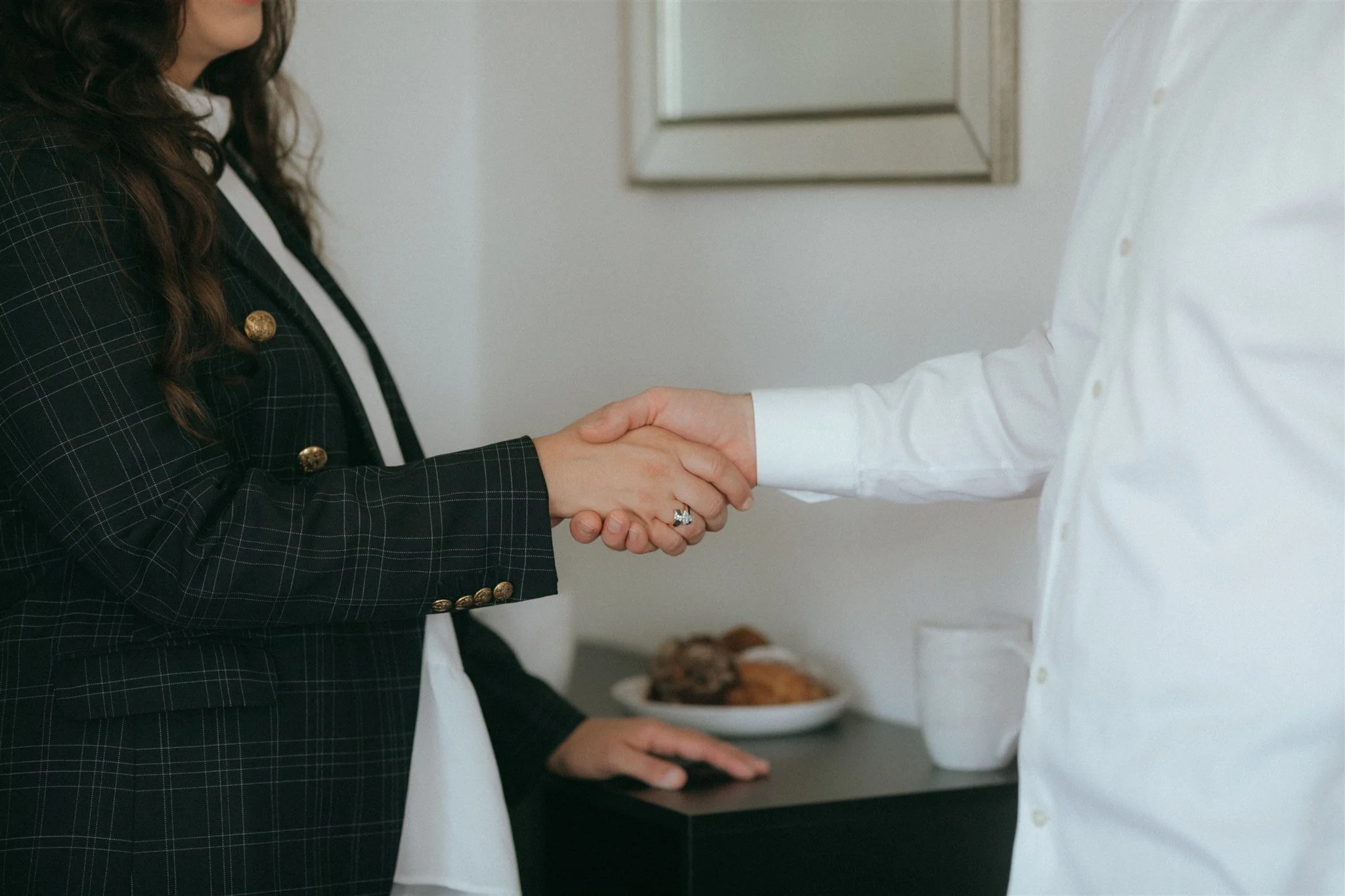Two people shaking hands, one wearing a dark plaid blazer with gold buttons, the other in a white shirt, in an indoor setting with a dark desk, a plate of baked goods, a glass of water, and a mirror on the wall.