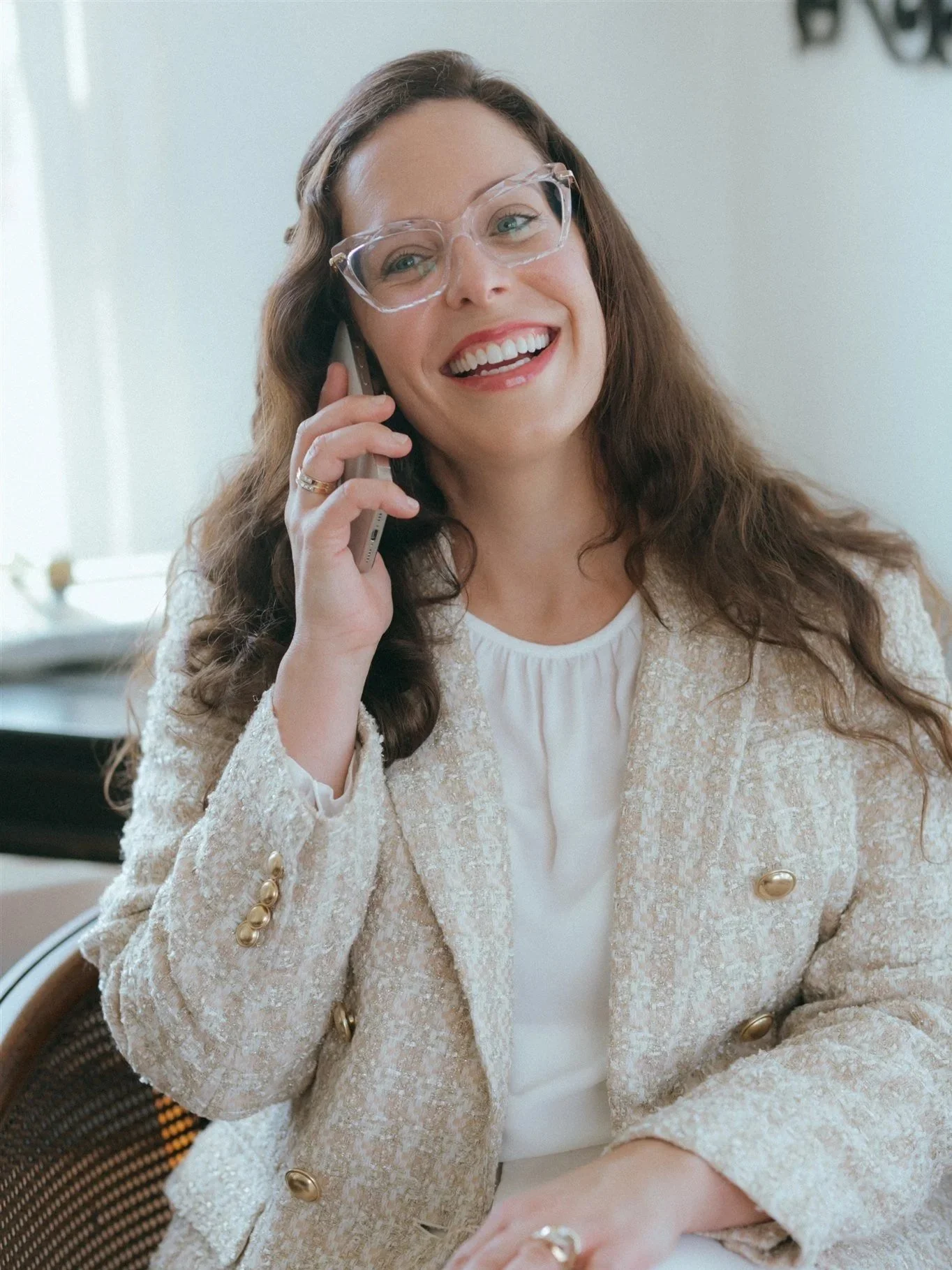 A woman with long, wavy hair smiling and talking on a cell phone, wearing clear glasses and a light-colored blazer.