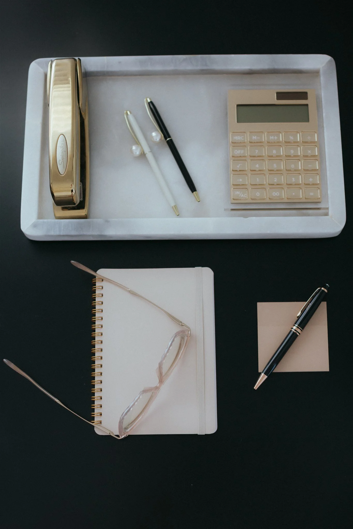 Desk organized with a gold stapler, two pens, a calculator, a notebook with a pair of glasses on top, a sticky note, and a small notepad on a black surface.