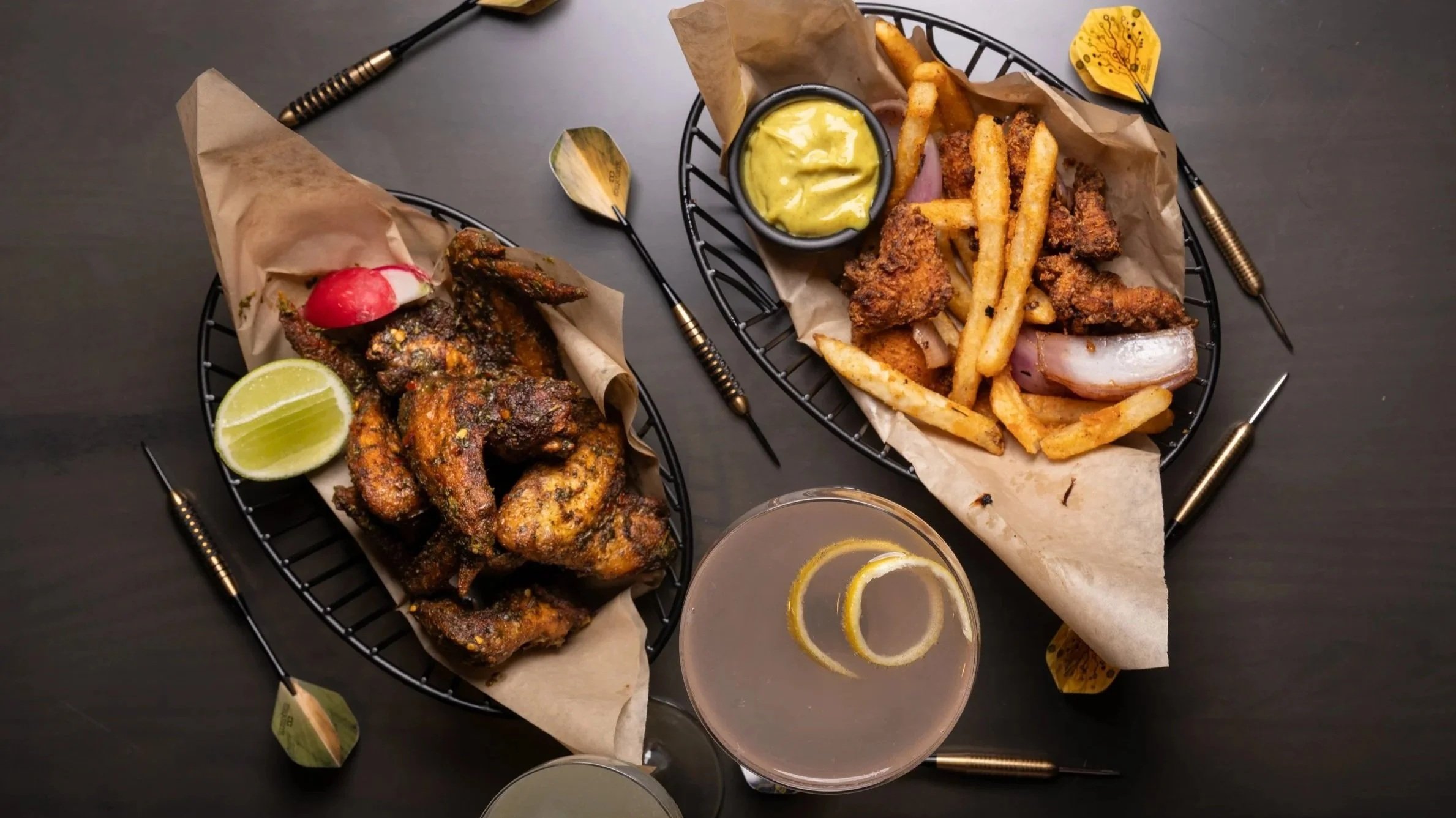 Image of a drink, darts, chicken wings and chicken bites with fries on a table.