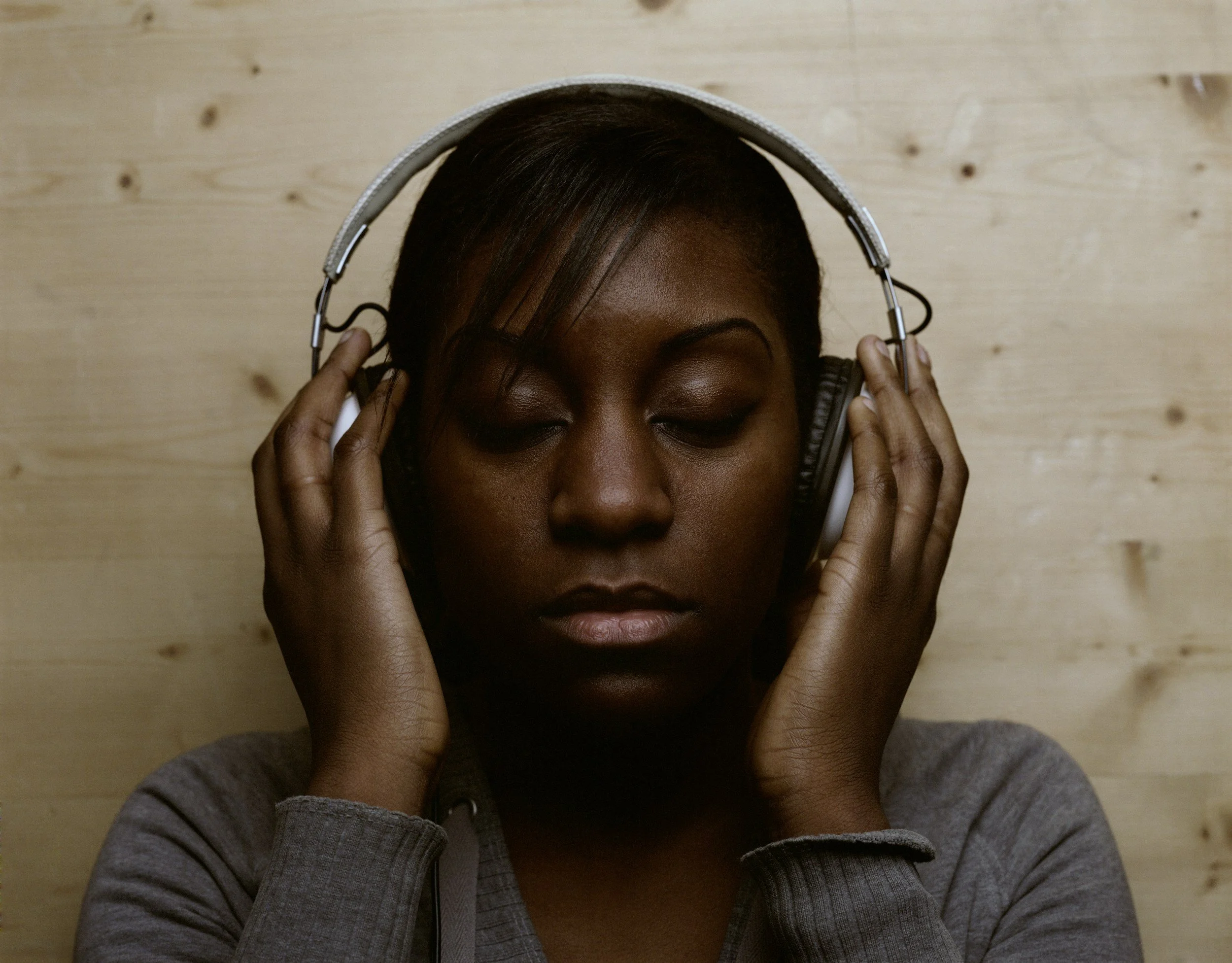 A woman with dark skin and closed eyes is listening to music with large over-ear headphones, against a wooden background.