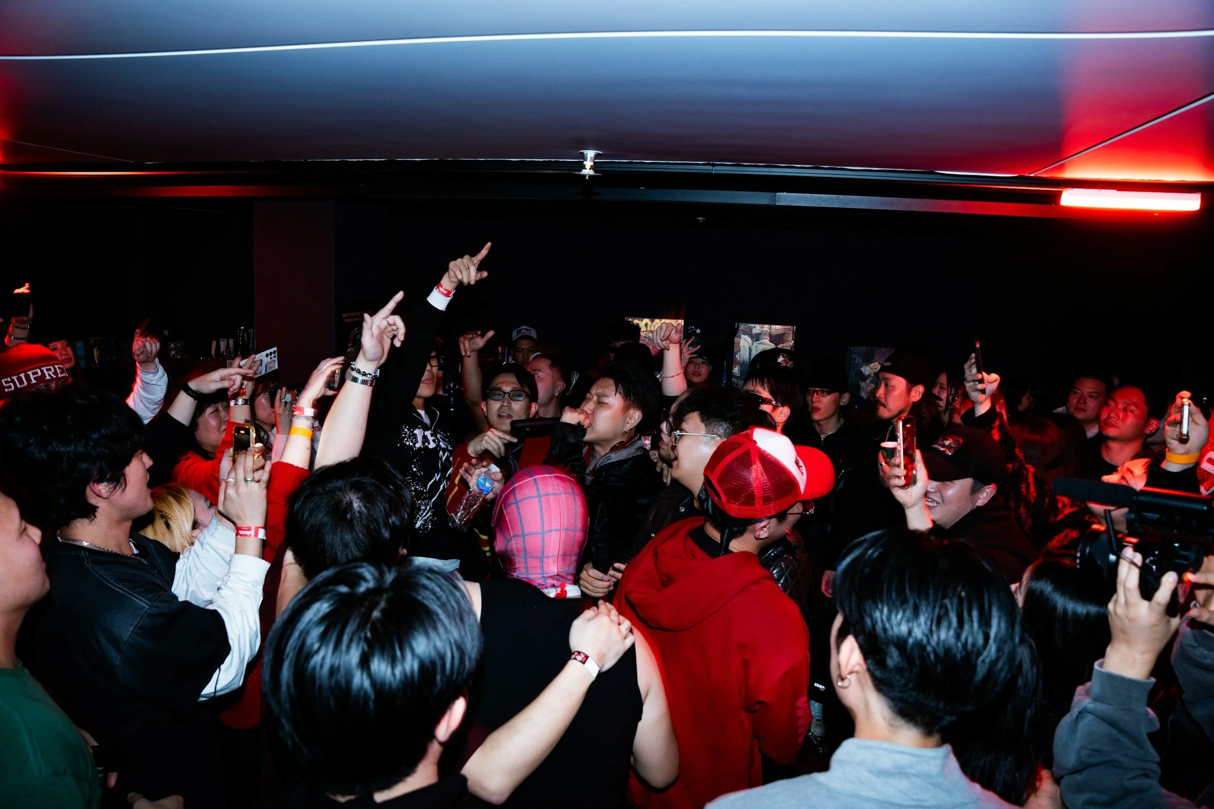 Crowd of people dancing and taking pictures in a dark, indoor nightclub with red lighting.