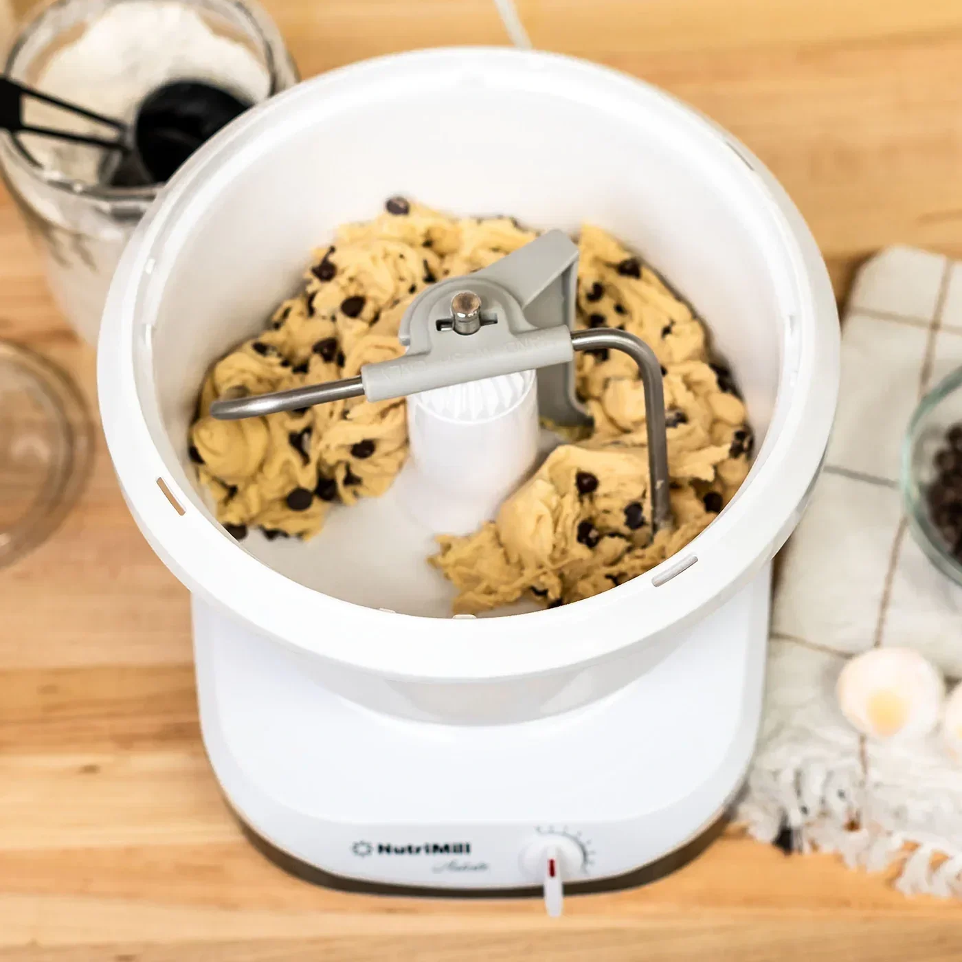 Chocolate chip cookie dough in a bread machine or mixer, with spoons and additional ingredients nearby on a wooden countertop.