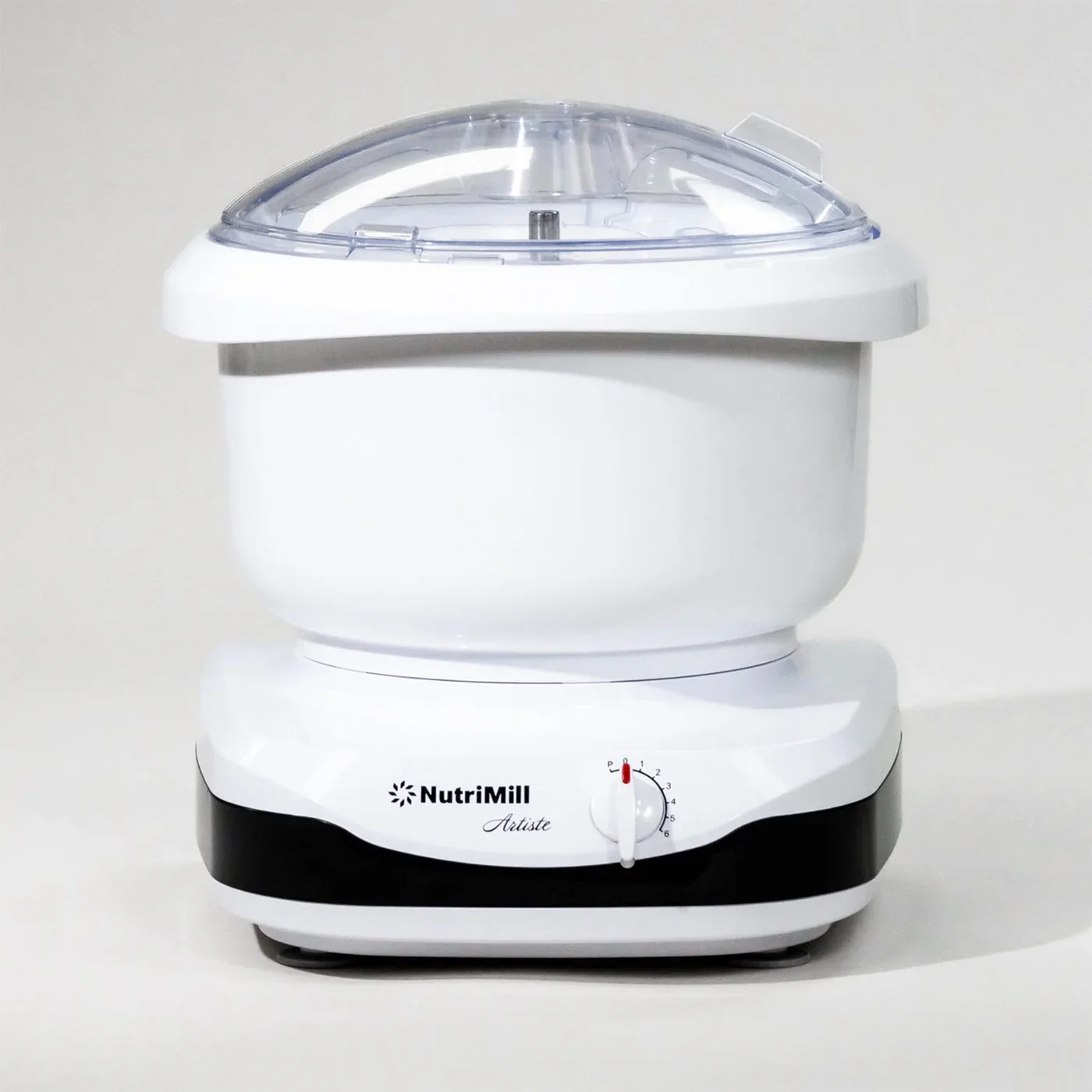 A white NutriMill Arista tabletop grain mill with a clear plastic lid, sitting on a white surface against a light-colored background.