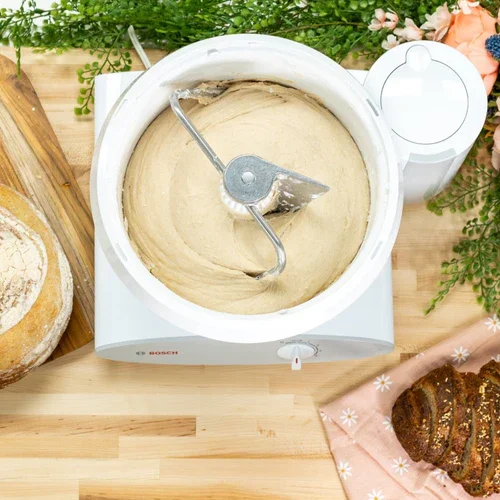 Ice cream maker machine with vanilla ice cream in a bowl, surrounded by bread, flowers, and a pink cloth.