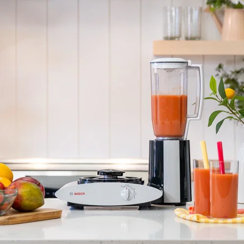 Blender filled with orange smoothie on kitchen countertop next to two glasses of smoothie with straws, kitchen plants and fruit in the background.