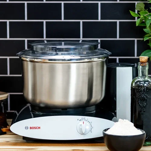 A Bosch stand mixer with a glass cover, placed on a wooden countertop in a kitchen, with a black bowl of flour in front and a black tiled wall in the background.