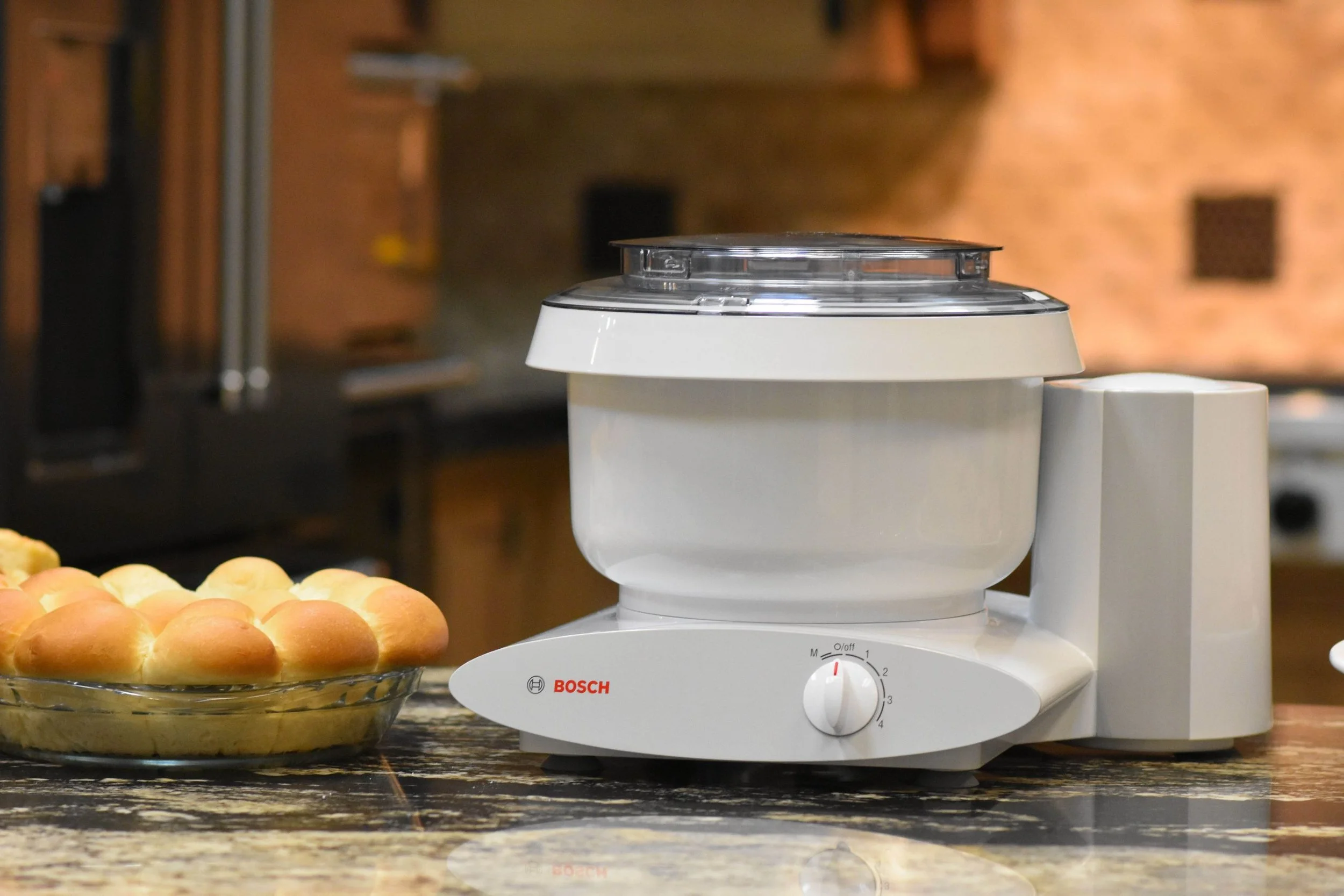 A white Bosch food processor on a kitchen countertop with a glass dish of bread rolls beside it.