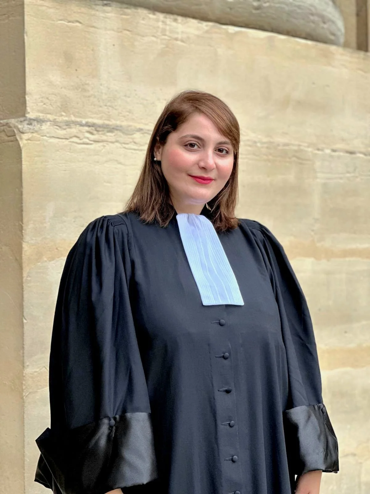 Une femme habillée d'une robe de juge ou d'officier en robe de cérémonie noire avec un col bleu clair, posant devant un mur en pierre. Elle a des cheveux bruns mi-longs et sourit légèrement.