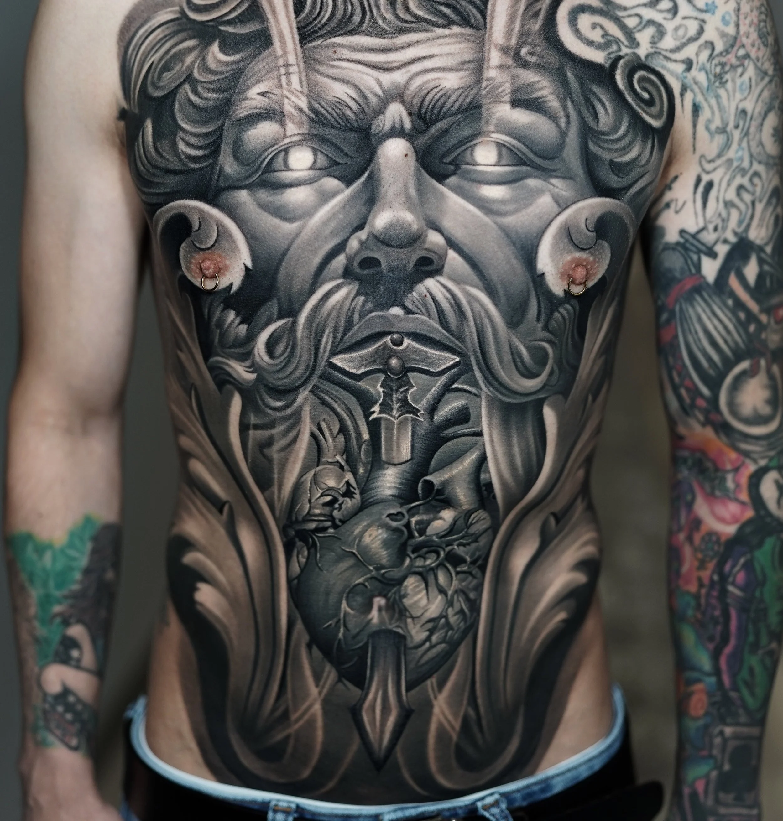 Black and gray tattoo of an old man's face with a beard and mustache, with a prominent nose and closed eyes, covering the chest and extending to the arms. The tattoo features detailed shading and intricate designs, including a heart at the center with a sword through it, surrounded by decorative elements.