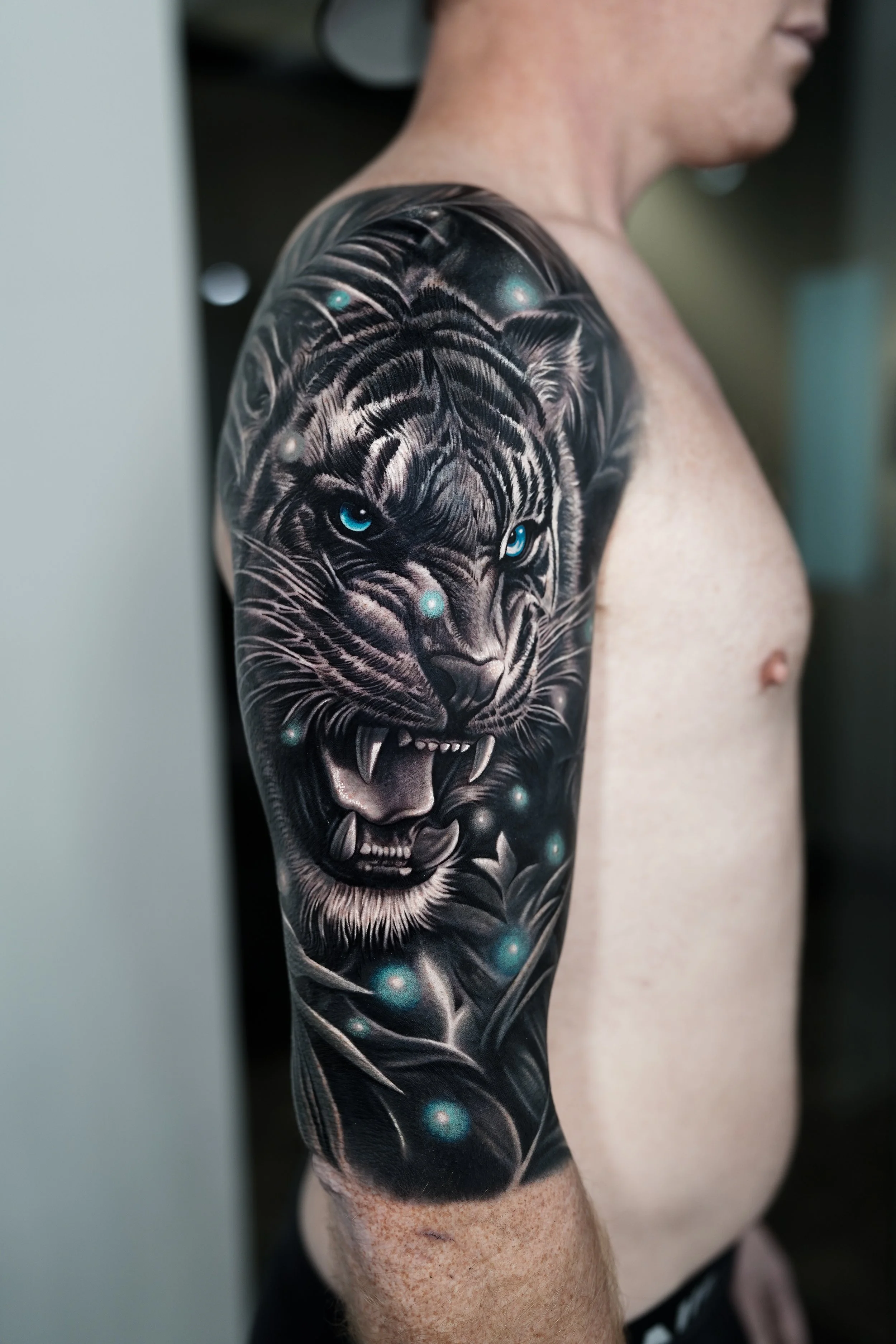 Close-up of a person's upper arm with a large, detailed black and gray tiger tattoo featuring blue eyes and glowing orbs.