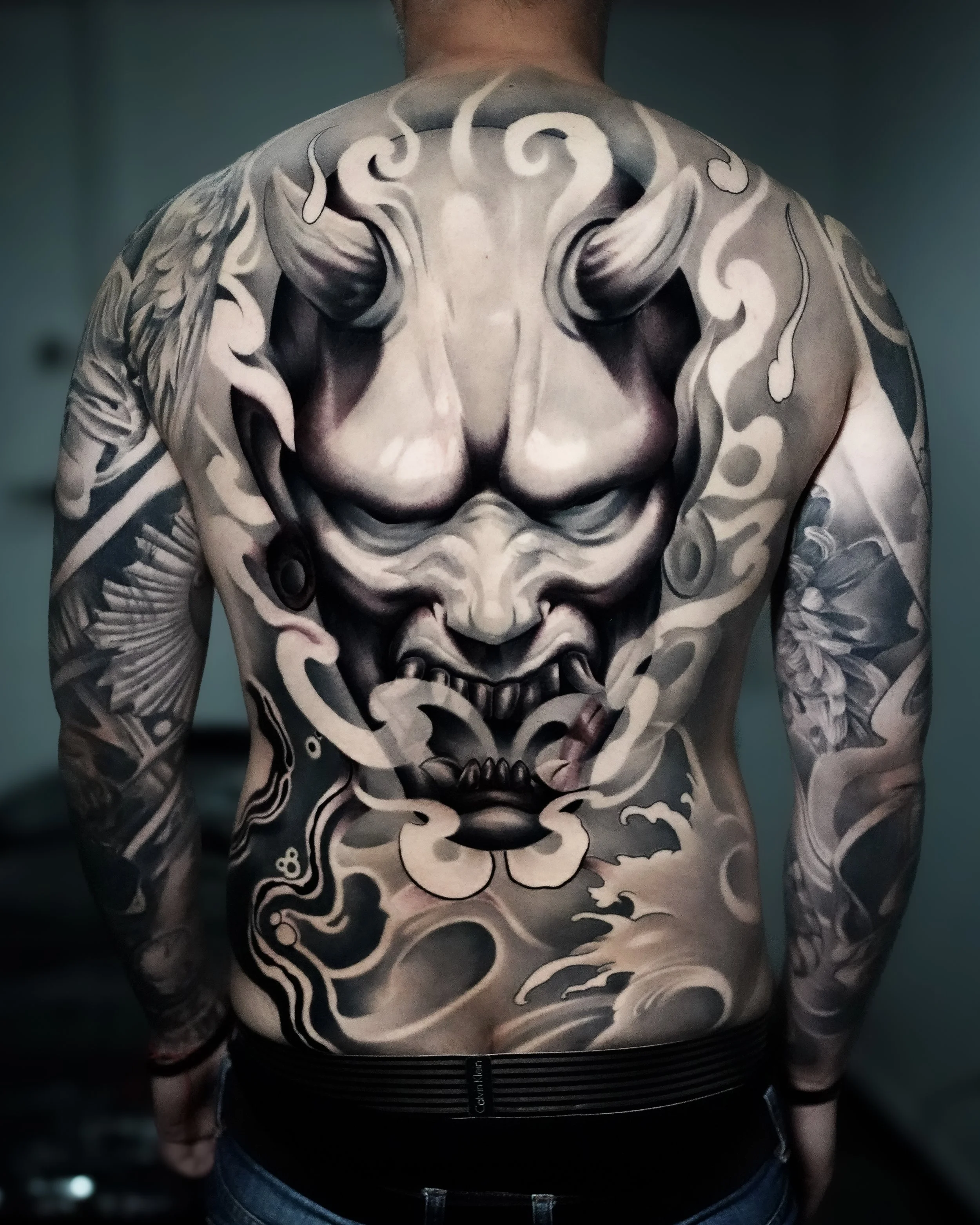 A person's back and arms with an intricate black and gray tiger tattoo design, featuring a fierce tiger face with sharp features and flowing elements.