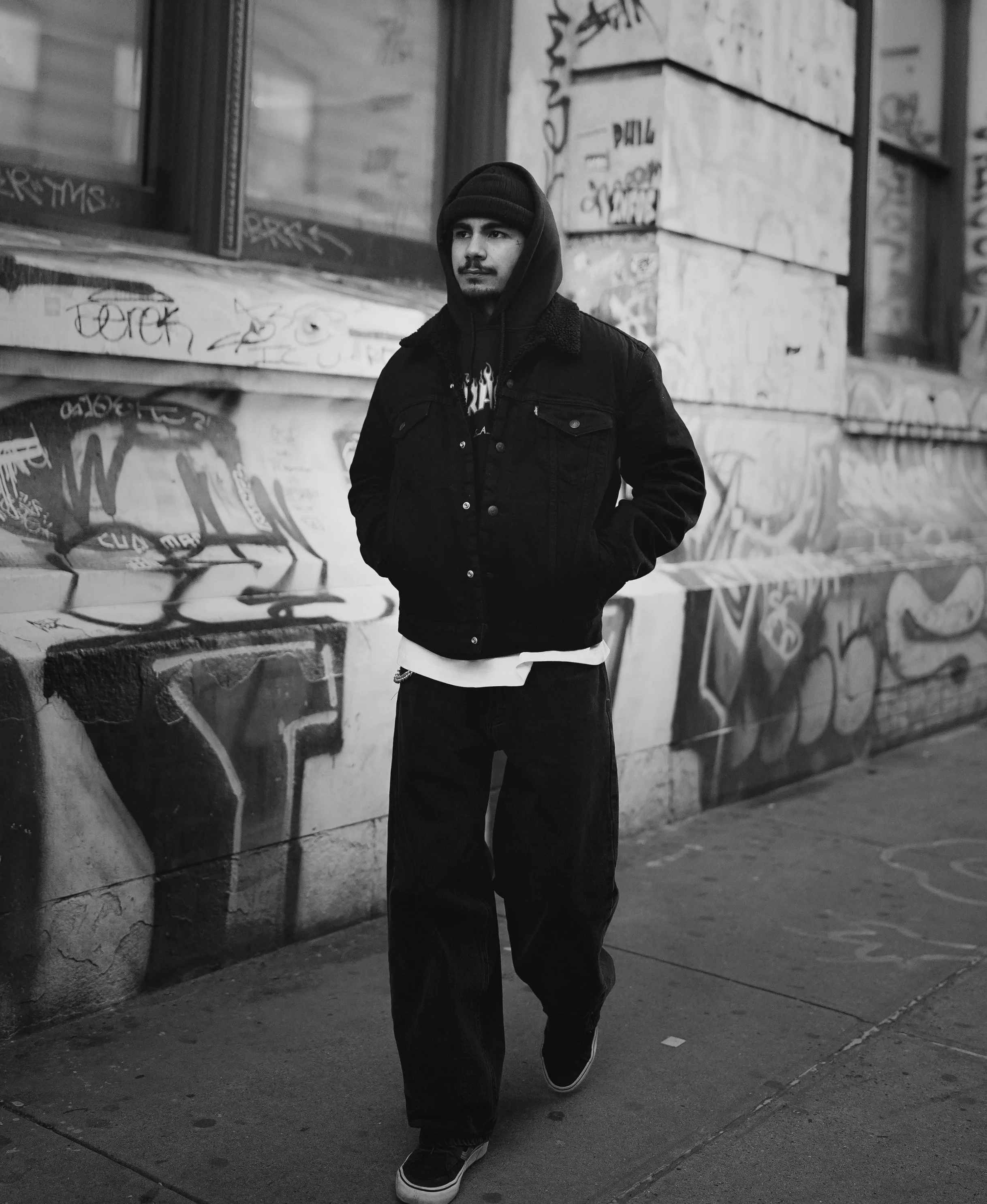 A young man wearing a black hoodie, black denim jacket, and baggy pants walking past a graffiti-covered wall in an urban setting.
