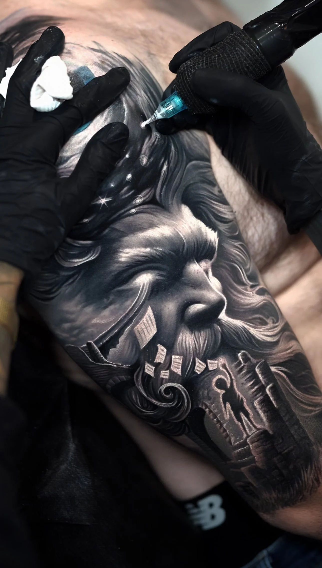 A person getting a black and gray tattoo of a bearded man's face, with long hair and a cosmic background, on their upper arm by a tattoo artist using a tattoo machine.