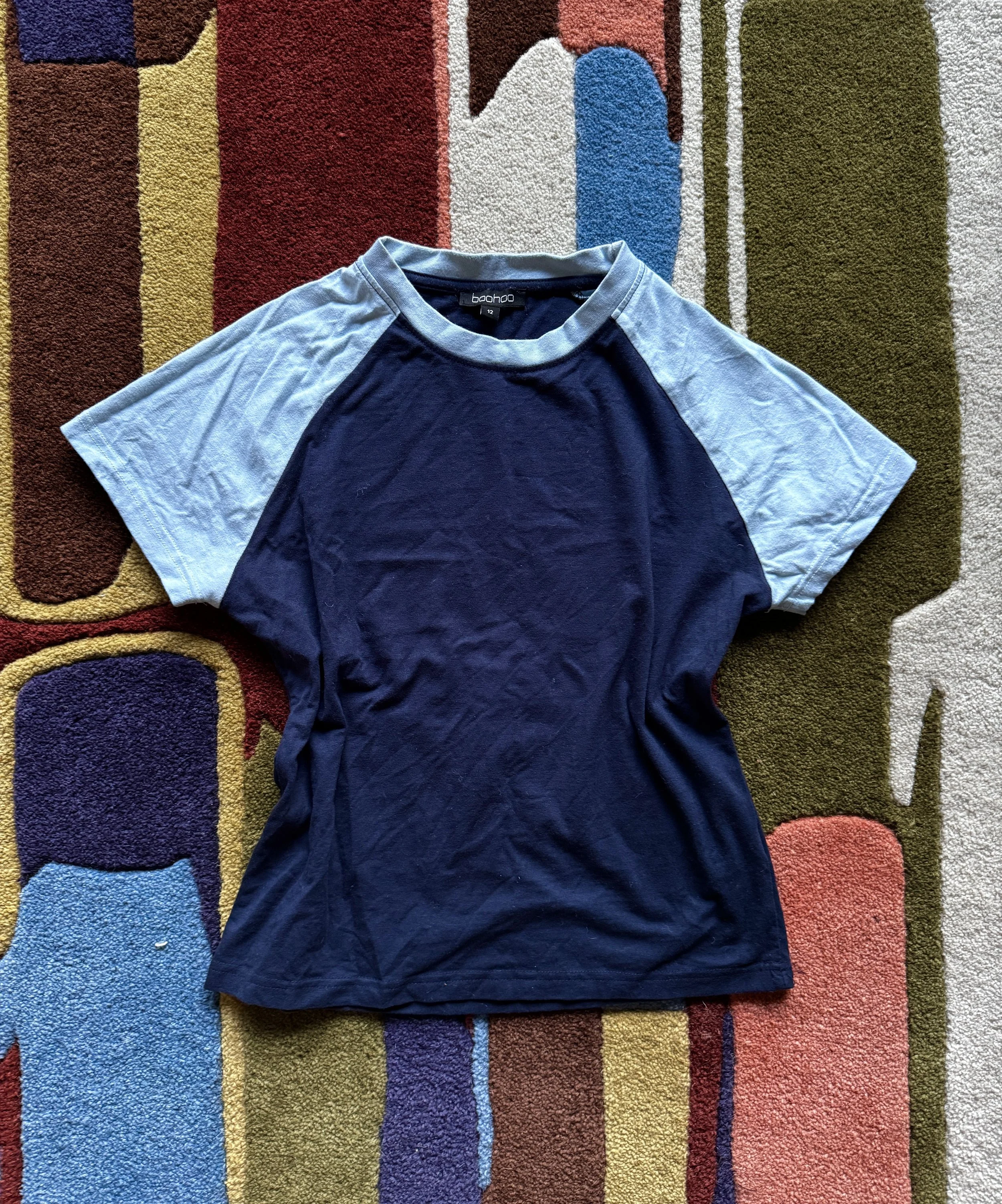 A blue and gray baseball-style T-shirt laid out flat on a colorful, abstract-patterned carpet.