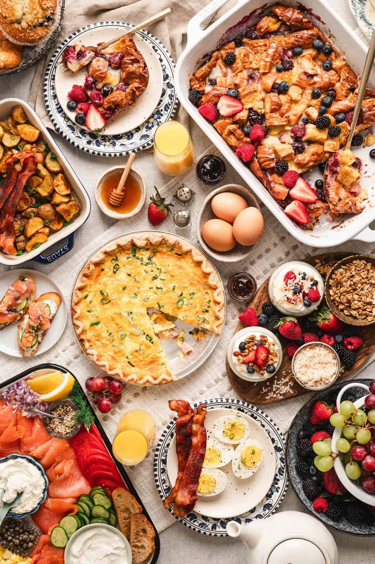 An assortment of breakfast and brunch foods including a fruit-topped casserole, quiche, eggs, bacon, smoked salmon platter, fresh berries, yogurt with granola, muffins, and glasses of orange juice on a table