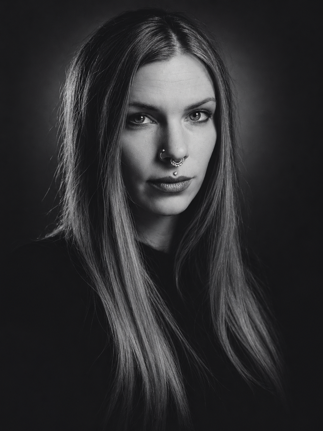 Black and white portrait of a young woman with long straight hair, tattoos, and multiple facial piercings, looking directly at the camera.