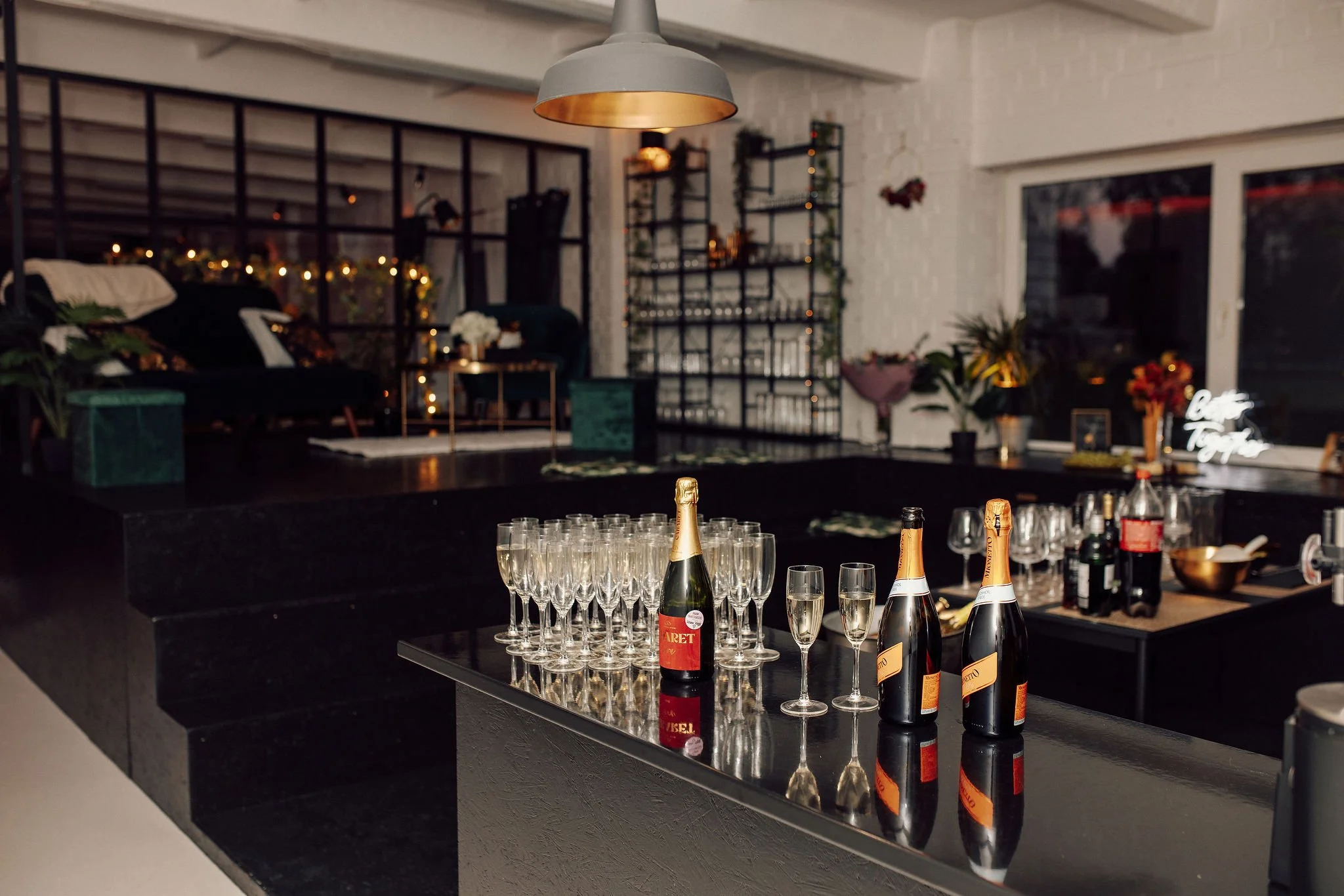 A bar area with bottles of sparkling wine and empty glasses on the counter. In the background, there is a cozy living space decorated with string lights, plants, and wall shelves, creating a warm and inviting atmosphere.