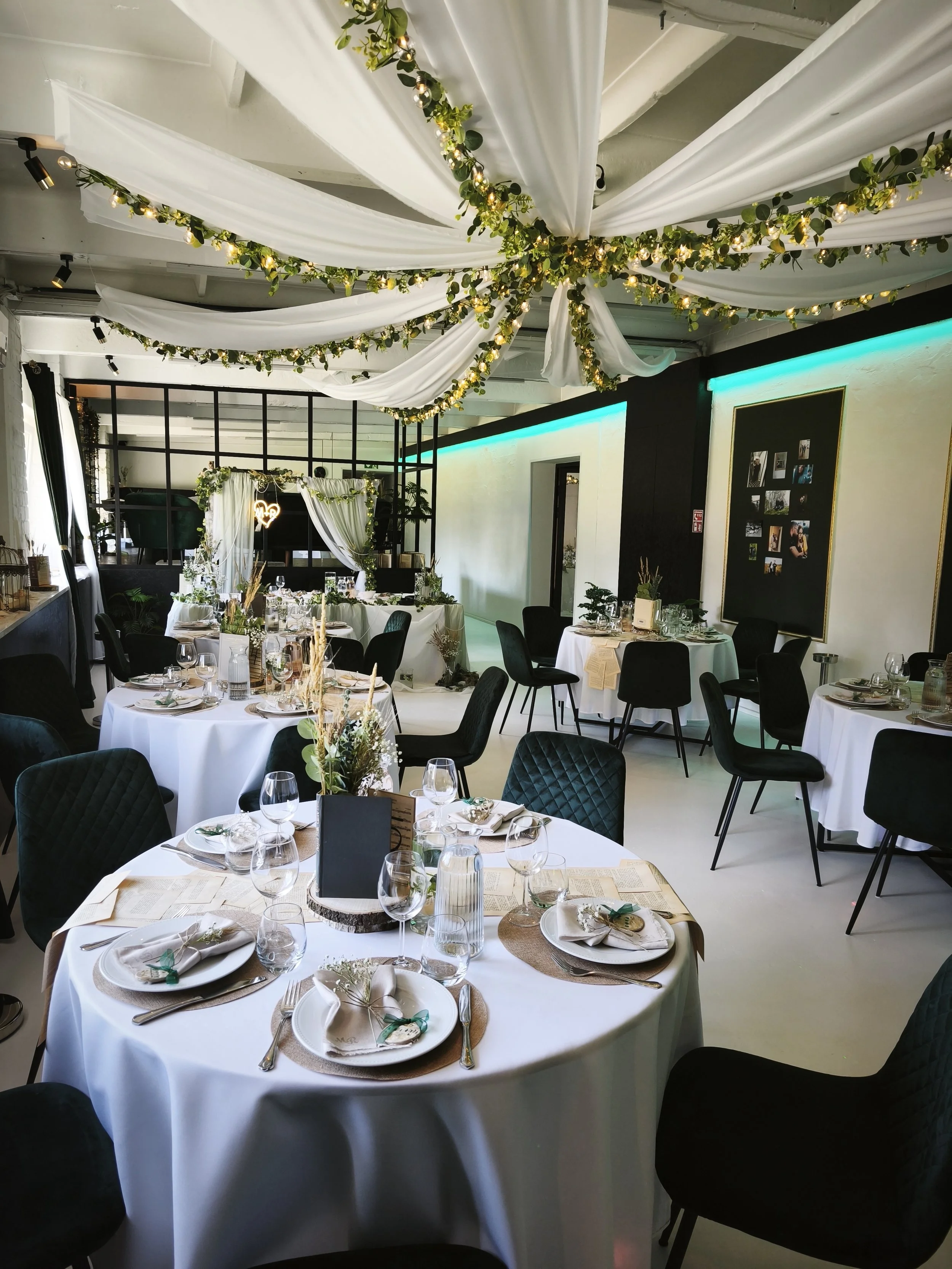 Wedding reception venue decorated with white drapes and string lights with greenery, featuring round tables with white tablecloths and elegant place settings.