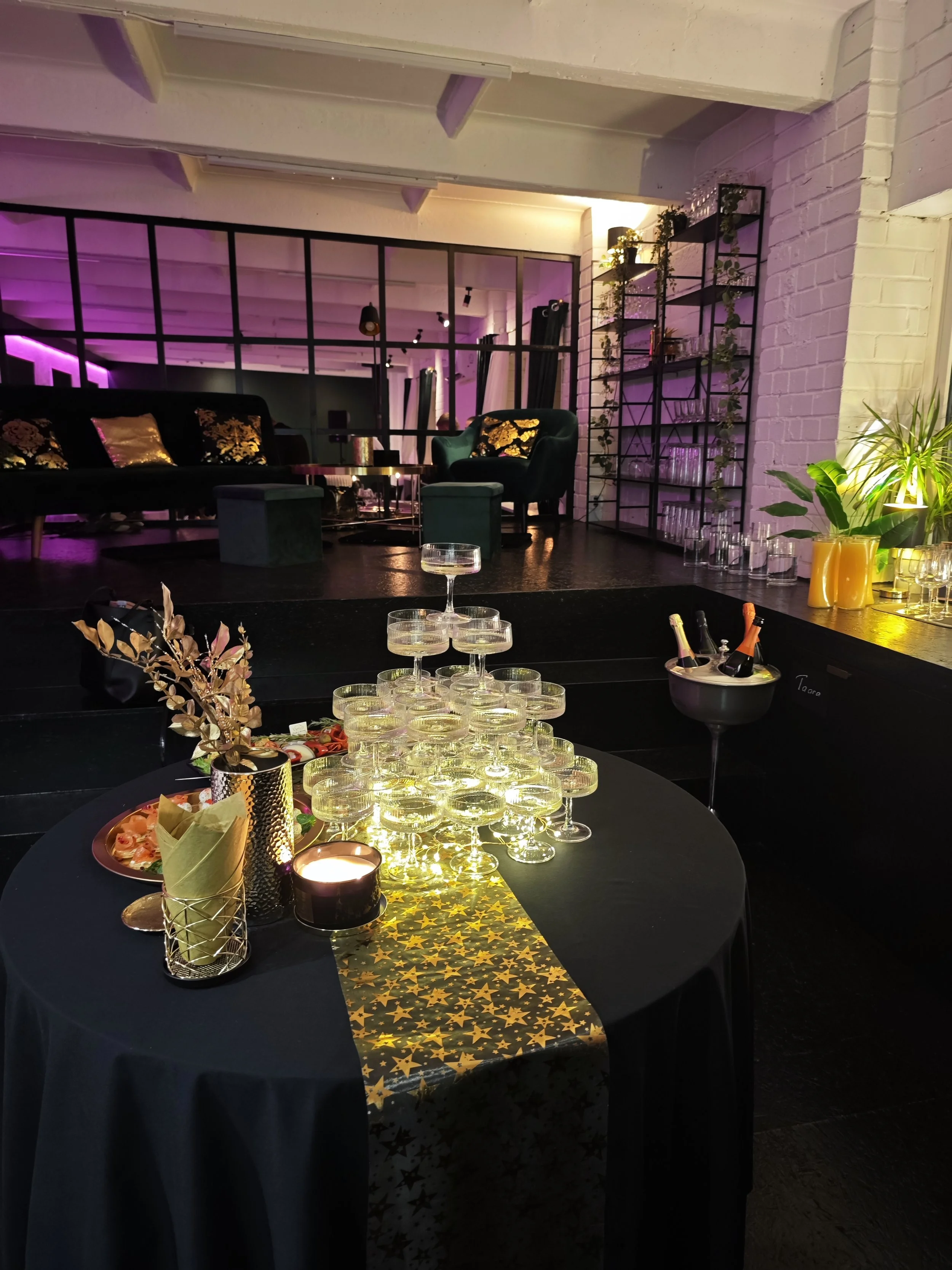 A decorated party table with champagne glasses, a candle, gold and black decorations, a gold star-patterned table runner, and a black tablecloth in a dimly lit room with purple, green, and yellow lighting.