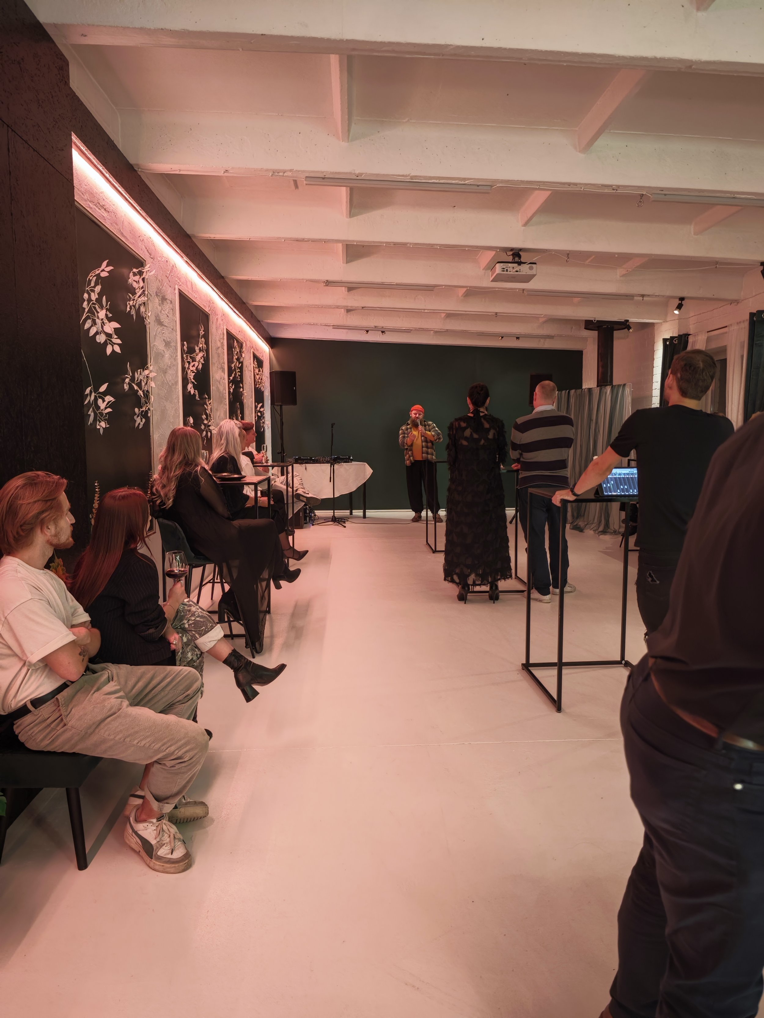 A group of people attending a small live performance or presentation in an indoor venue with a minimalist decor, featuring a performer on stage with a microphone and audience members seated or standing, some with drinks.