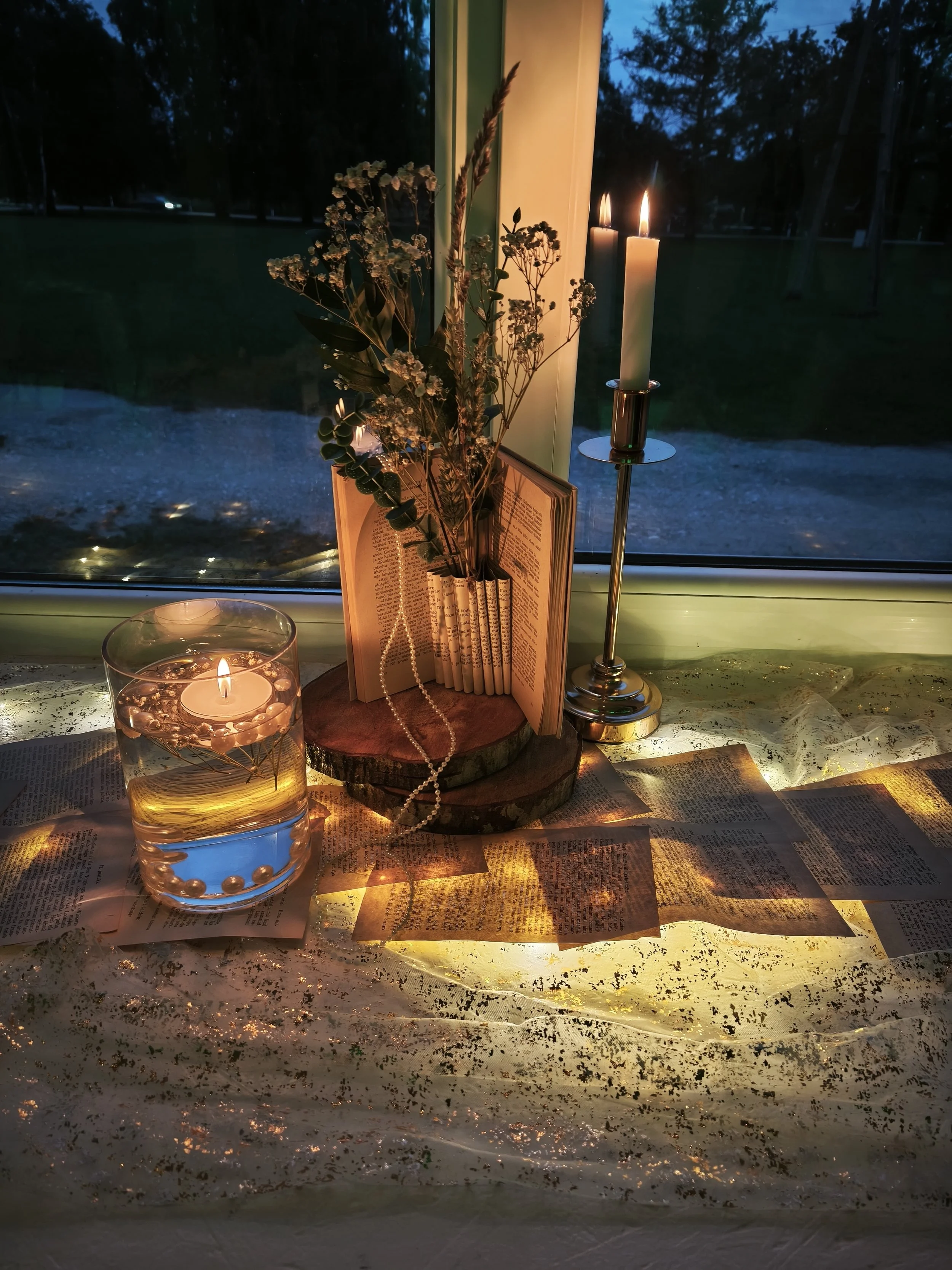A table decorated with candles, flowers, books, rosary beads, and scattered gold glitter, set in front of a window with a outdoor view at dusk.