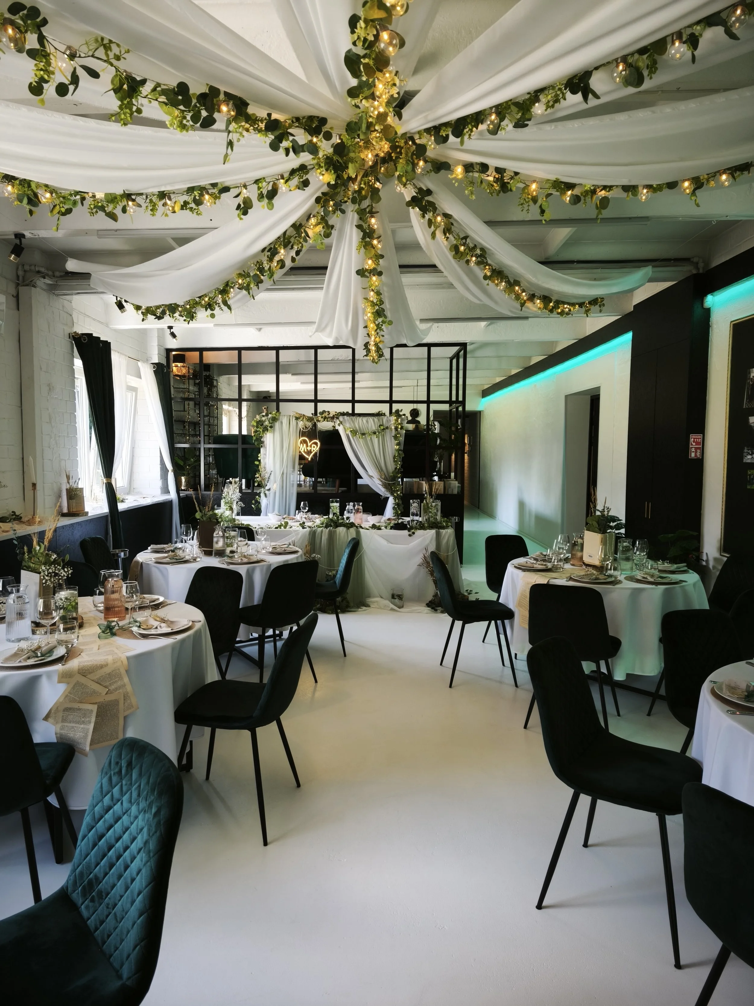 Festive event space decorated with white draped fabric and greenery with string lights on the ceiling, set with round tables, black chairs, and a decorated head table with floral arrangements.