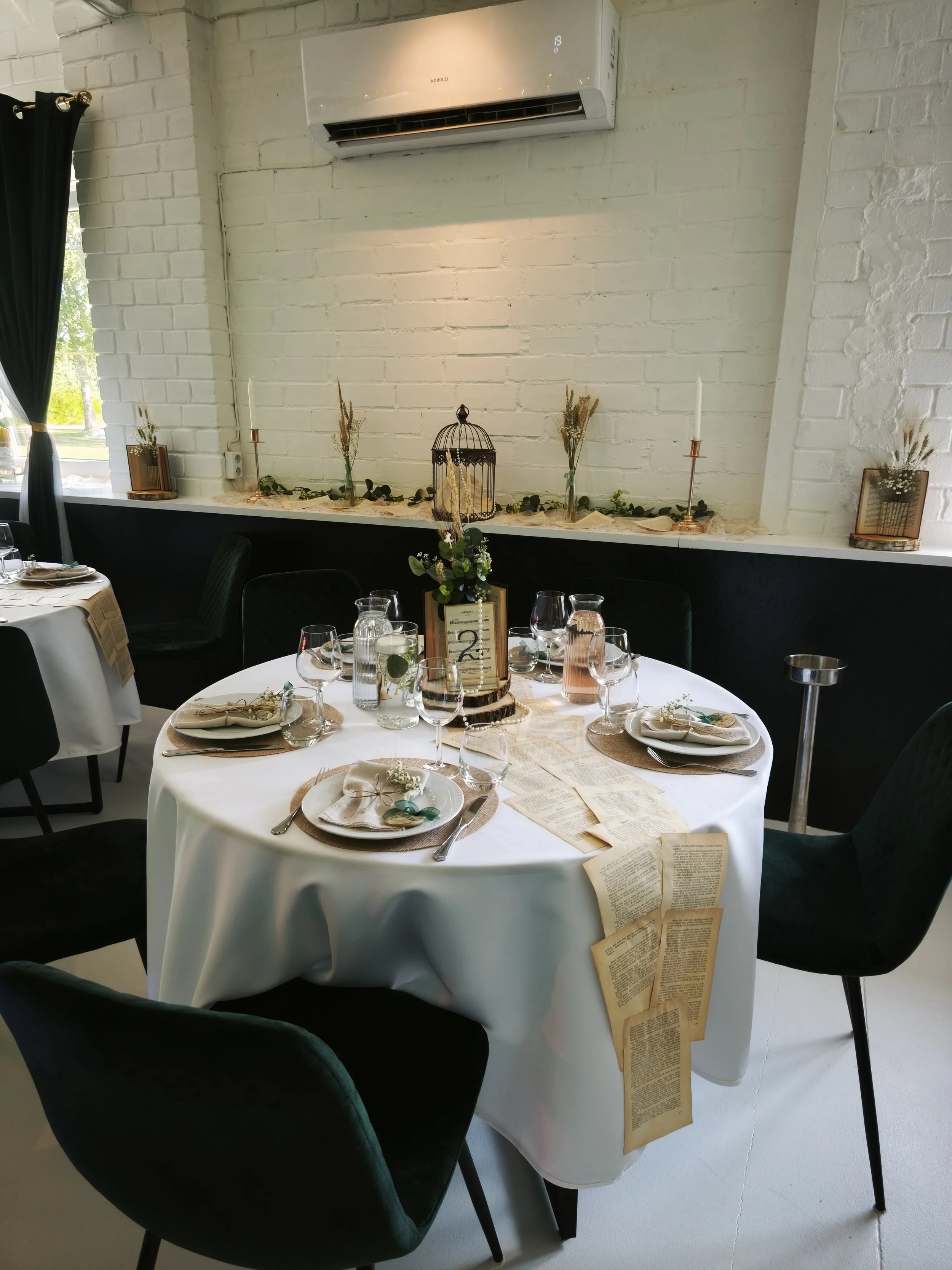 Round table set for a meal with a white tablecloth, glassware, plates, and napkins with small floral decorations, in a decorated event space with a white brick wall, black chairs, and a display with candles, greenery, and some papers.