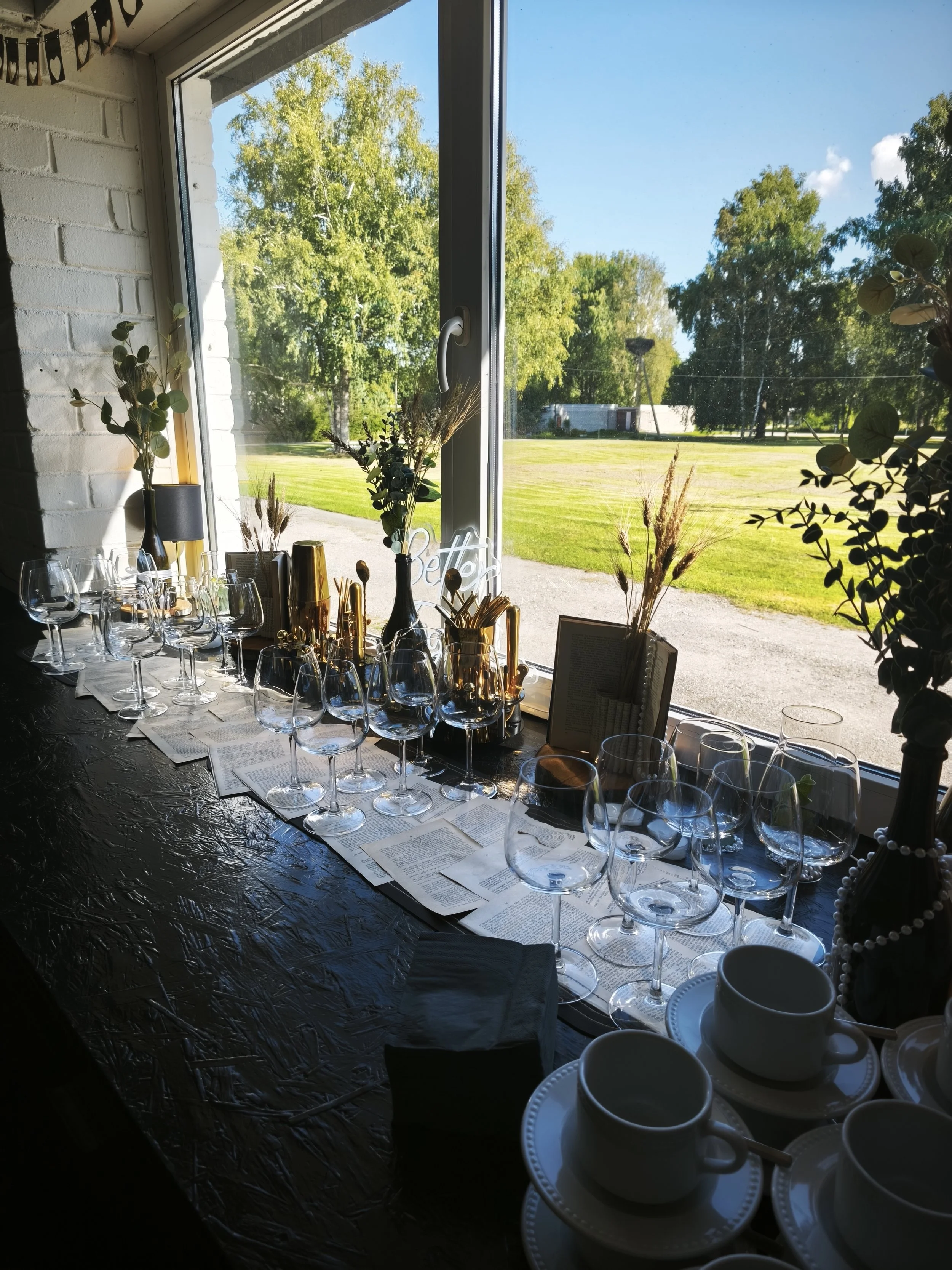 A window view of a grassy outdoor area with trees and a clear blue sky. Inside, a table is set with multiple wine glasses, cups, and decorative items, including dried flowers and greenery, with sunlight illuminating the scene.