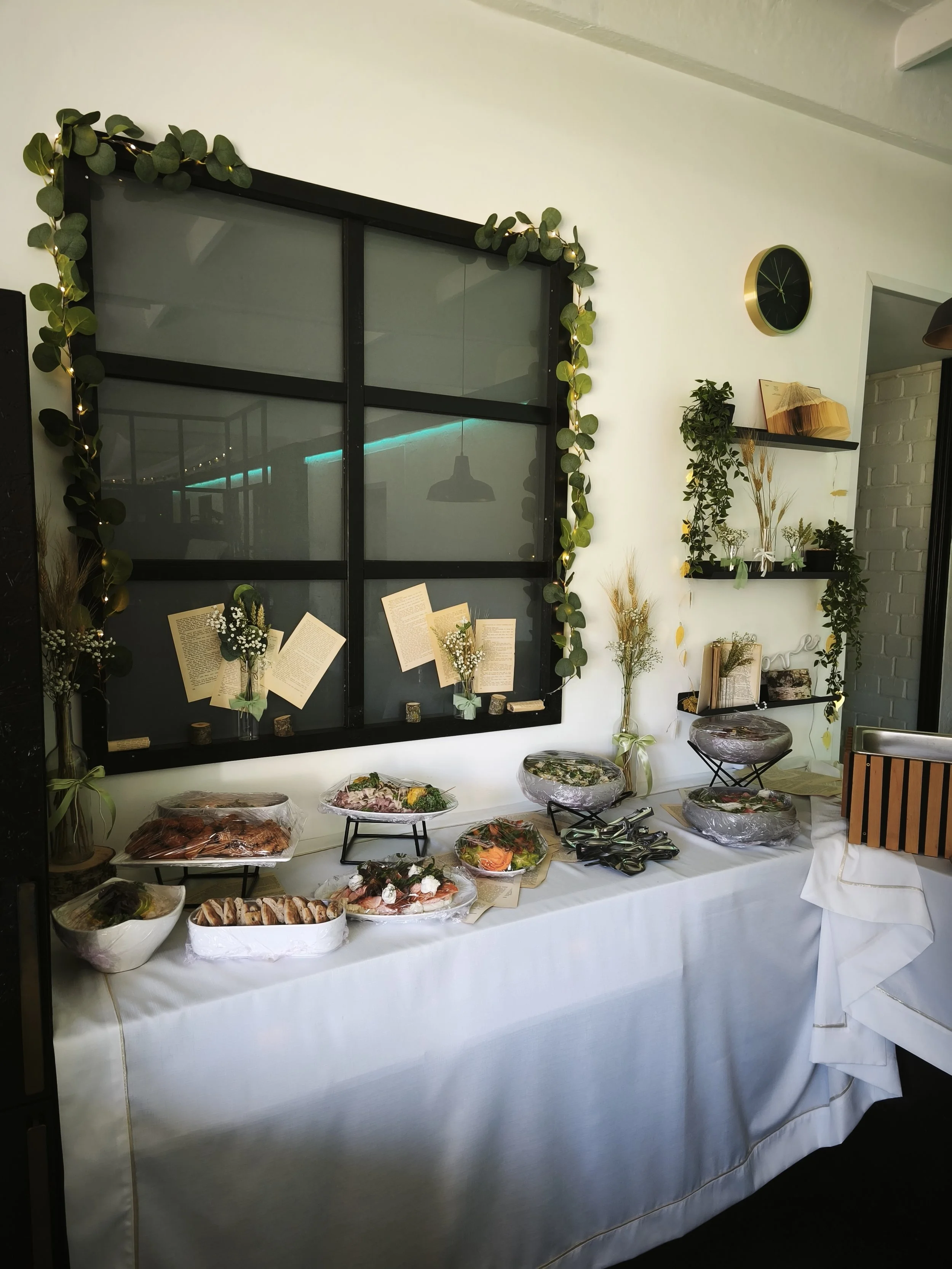 Table with a white tablecloth holding various dishes, including baked goods, fruits, and salads, in clear plastic and ceramic bowls, set against a decorated wall with shelves, greenery, and a clock.