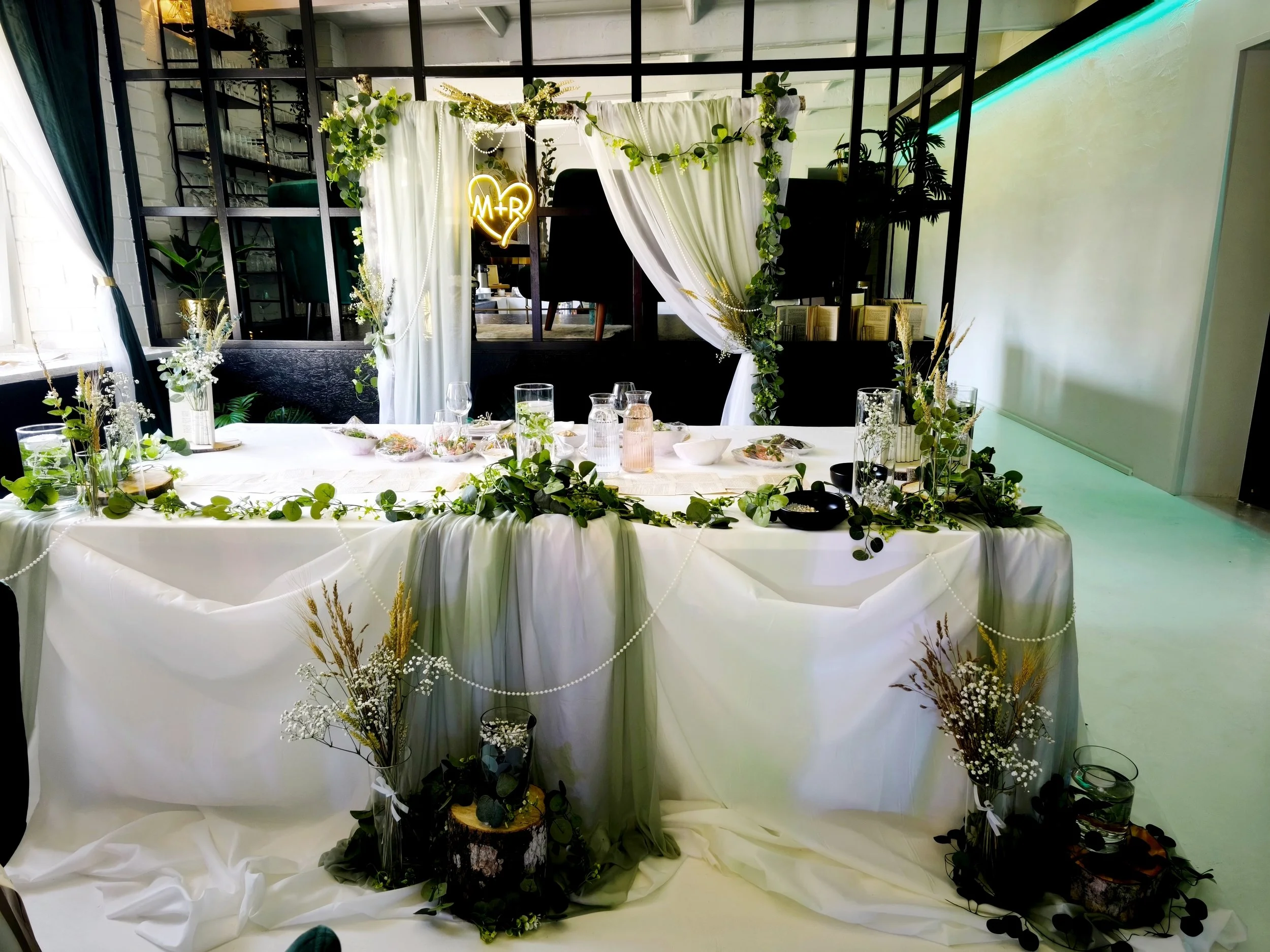 A decorated table with white cloths and greenery, arranged for a celebration or event. There are glasses, bowls, and small floral arrangements on the table. Behind the table, a backdrop of white drapes decorated with greenery and a yellow neon heart 