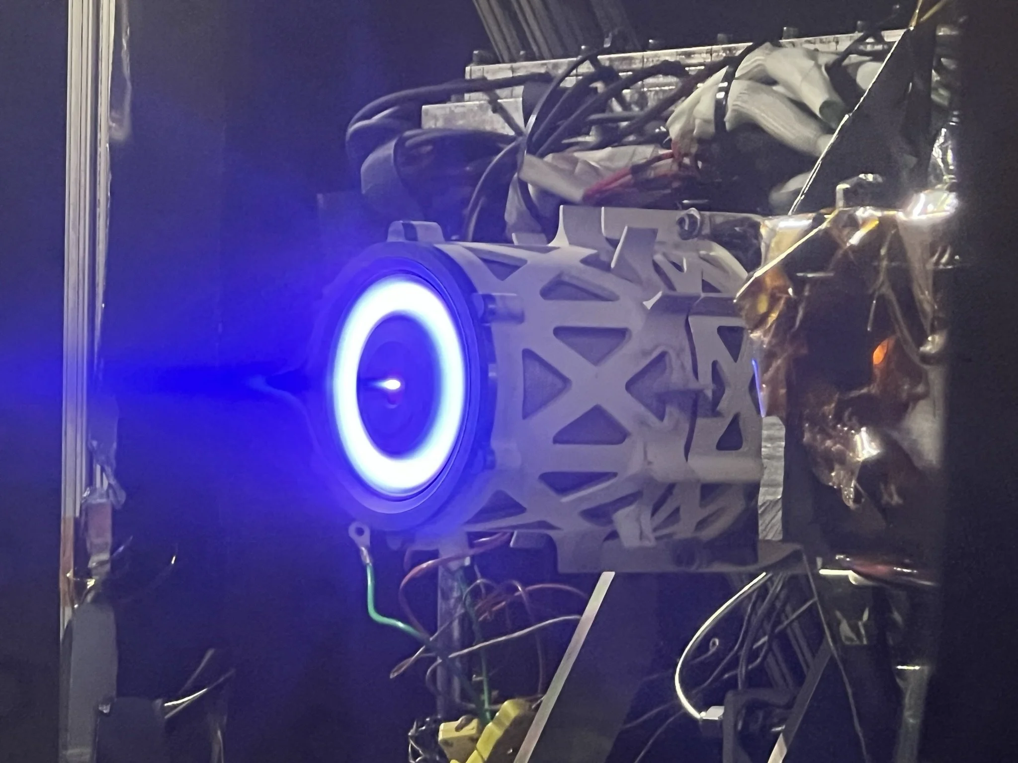 A close-up of a mechanical or electronic device with a white structural component, black wires, and a glowing ring of blue light, all set against a dark background.