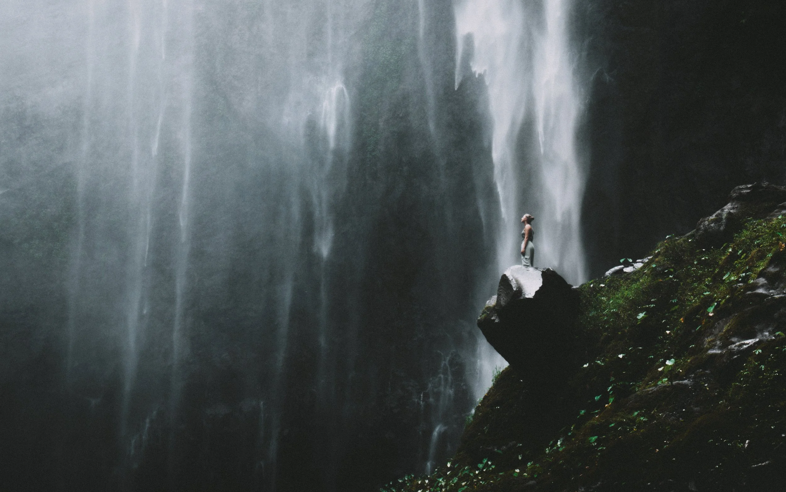 A woman standing on a large rock in front of a cascading waterfall with mist and greenery