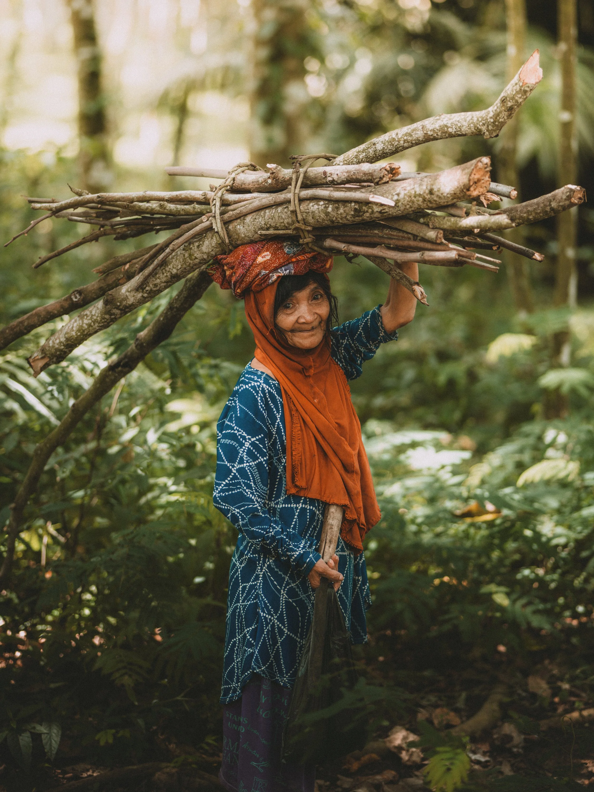 An elderly woman hiking through a forest with a large bundle of firewood balanced on her head and holding a stick.