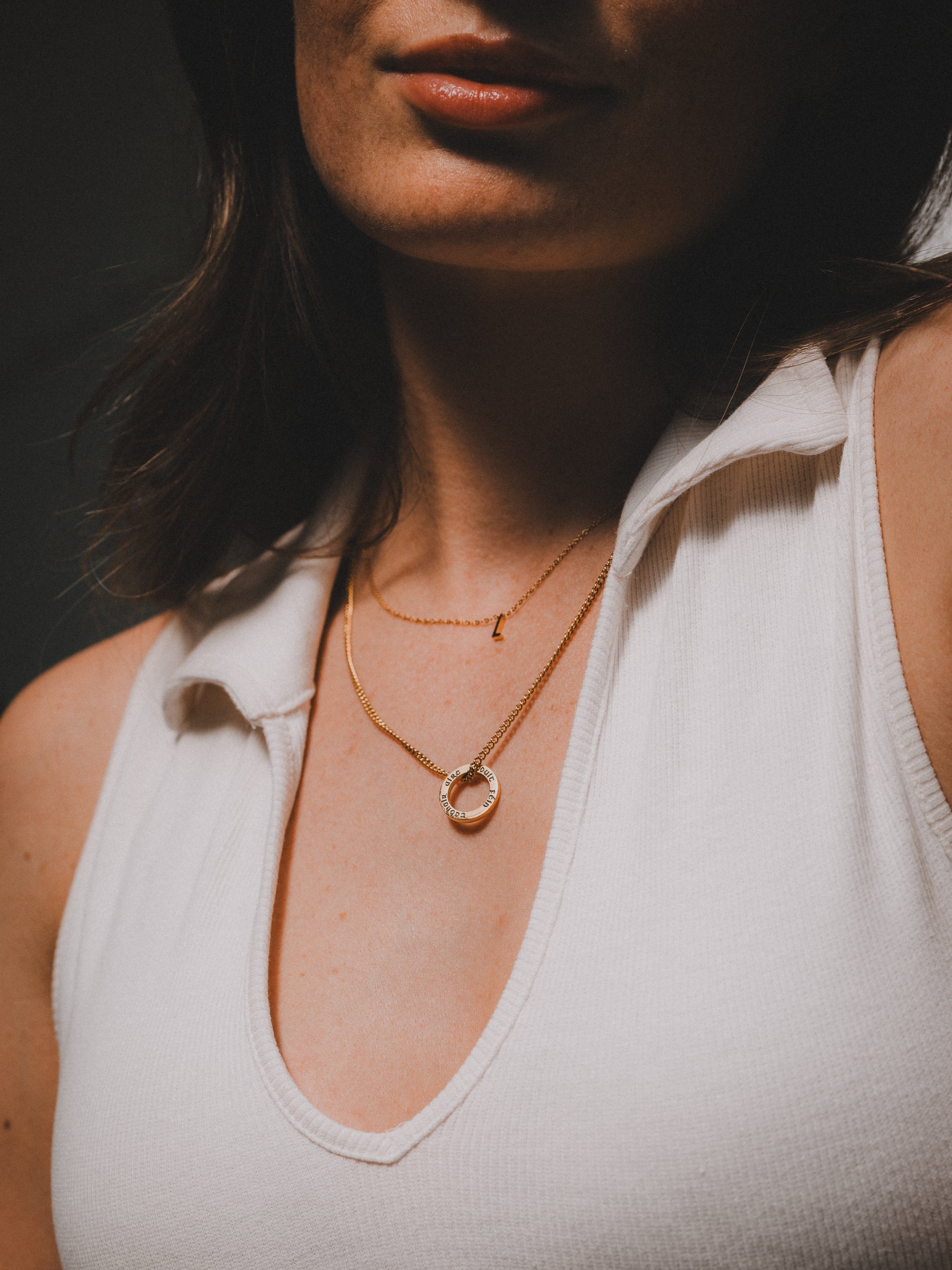 Close-up of a woman wearing layered gold necklaces, one with a small pendant and another with a circular pendant with engraved text, dressed in a white sleeveless top.