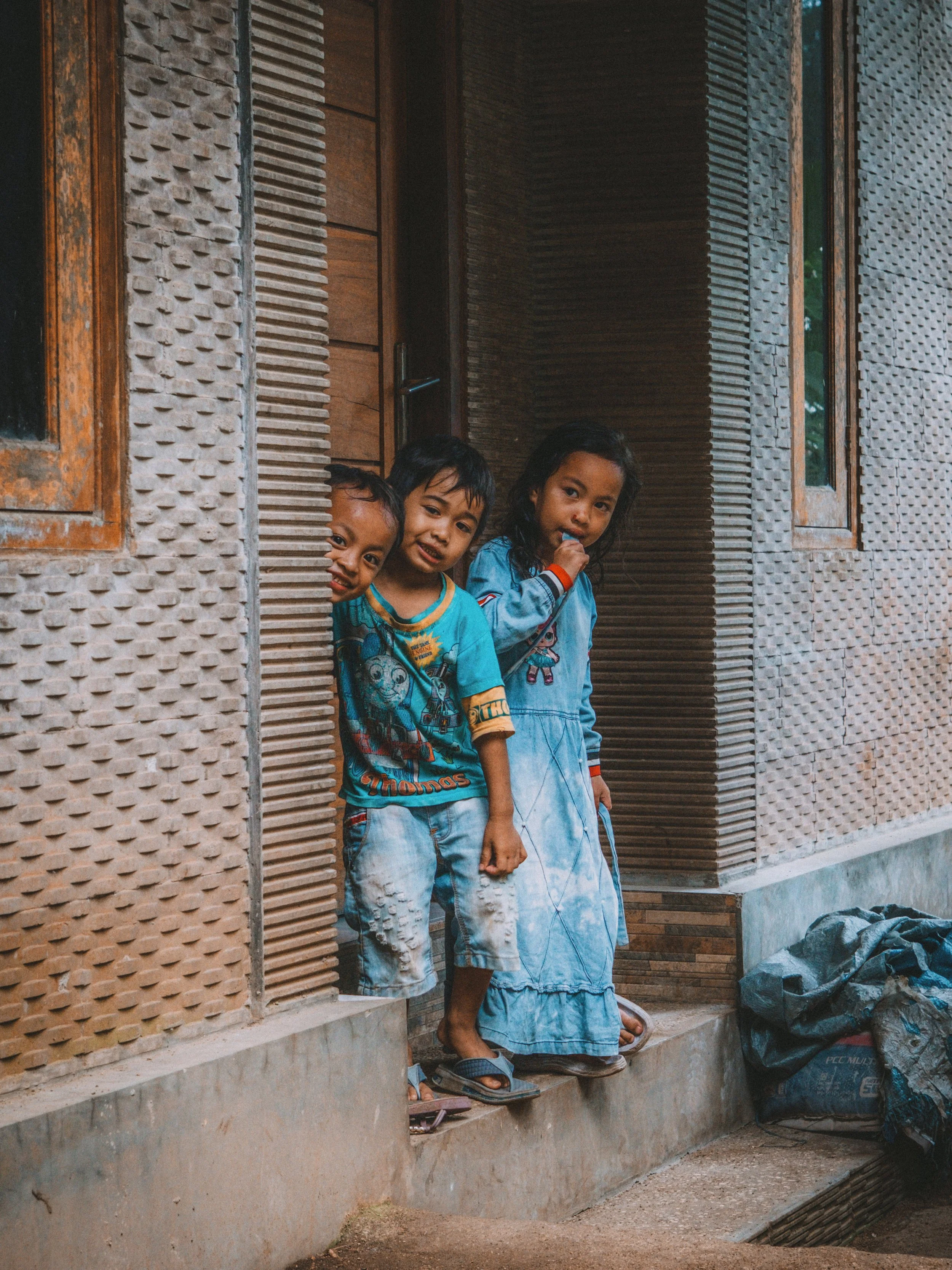 Three children peeking out from a doorway of a rustic house.