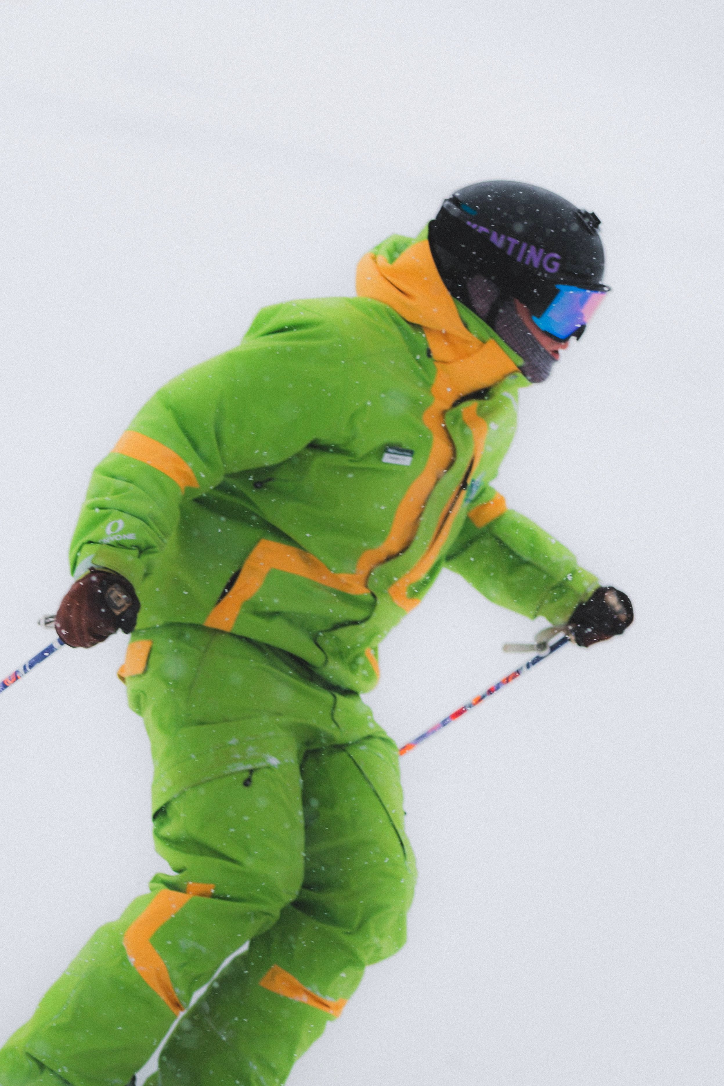 A person dressed in bright green and orange ski gear, wearing a black helmet and goggles, skiing in a snowy environment.