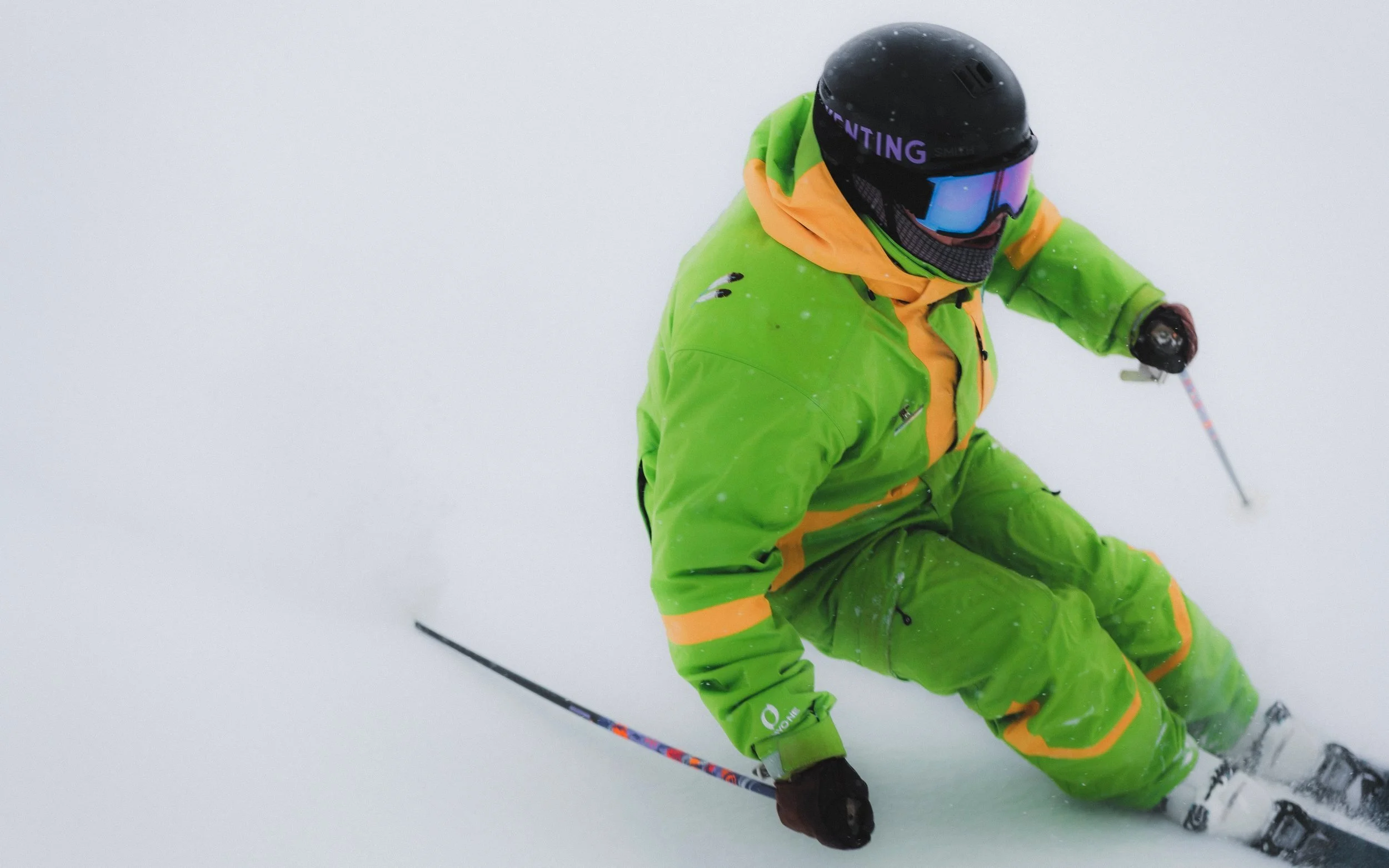 Skier in green and orange gear skiing on snowy slope with snow spray and foggy background.