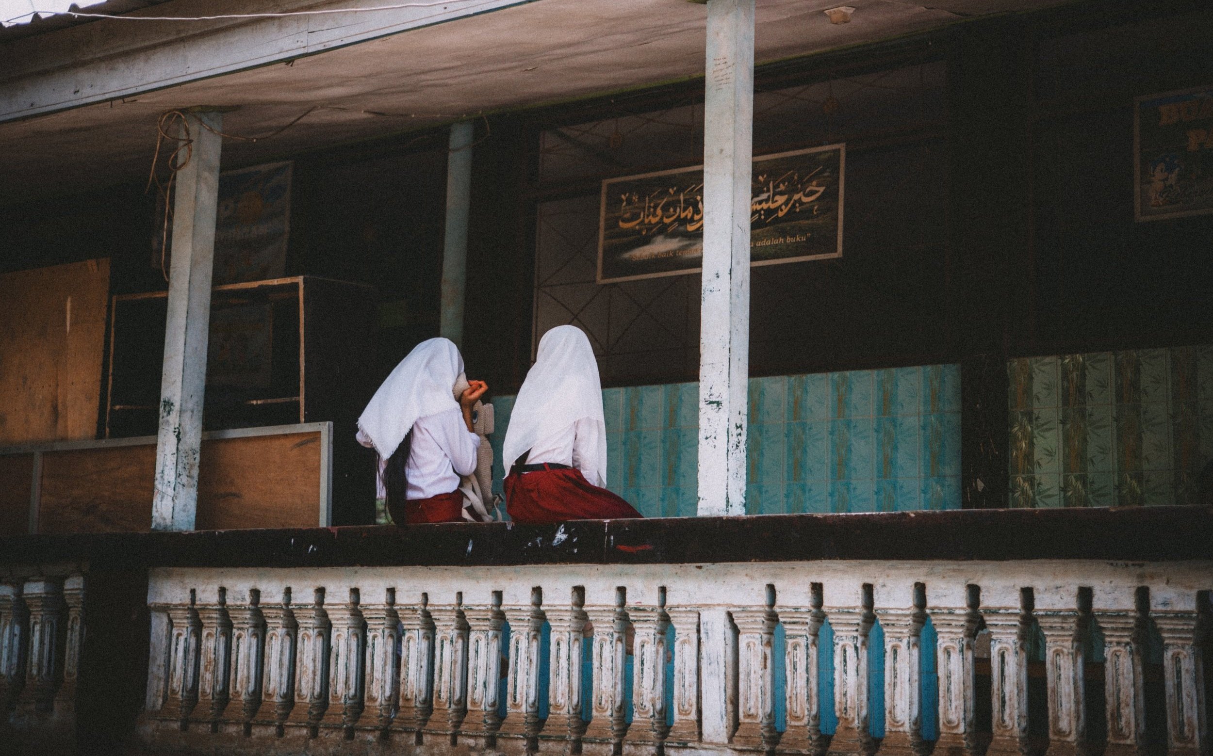 Two young girls wearing white headscarves and red skirts sitting on a ledge in an outdoor area, talking to each other, with a dark background and signs with foreign script behind them.