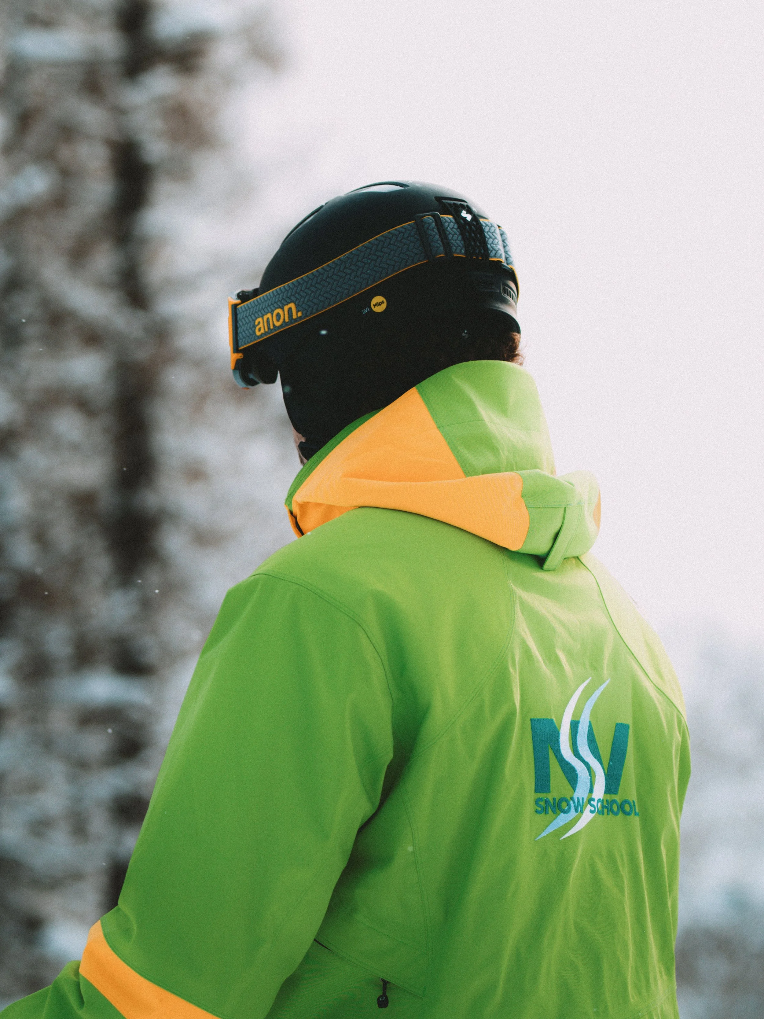 A snowboarding instructor wearing a black helmet with a yellow and blue band, a bright green and orange jacket with 'Snow School' logo, standing outdoors in a snowy environment.