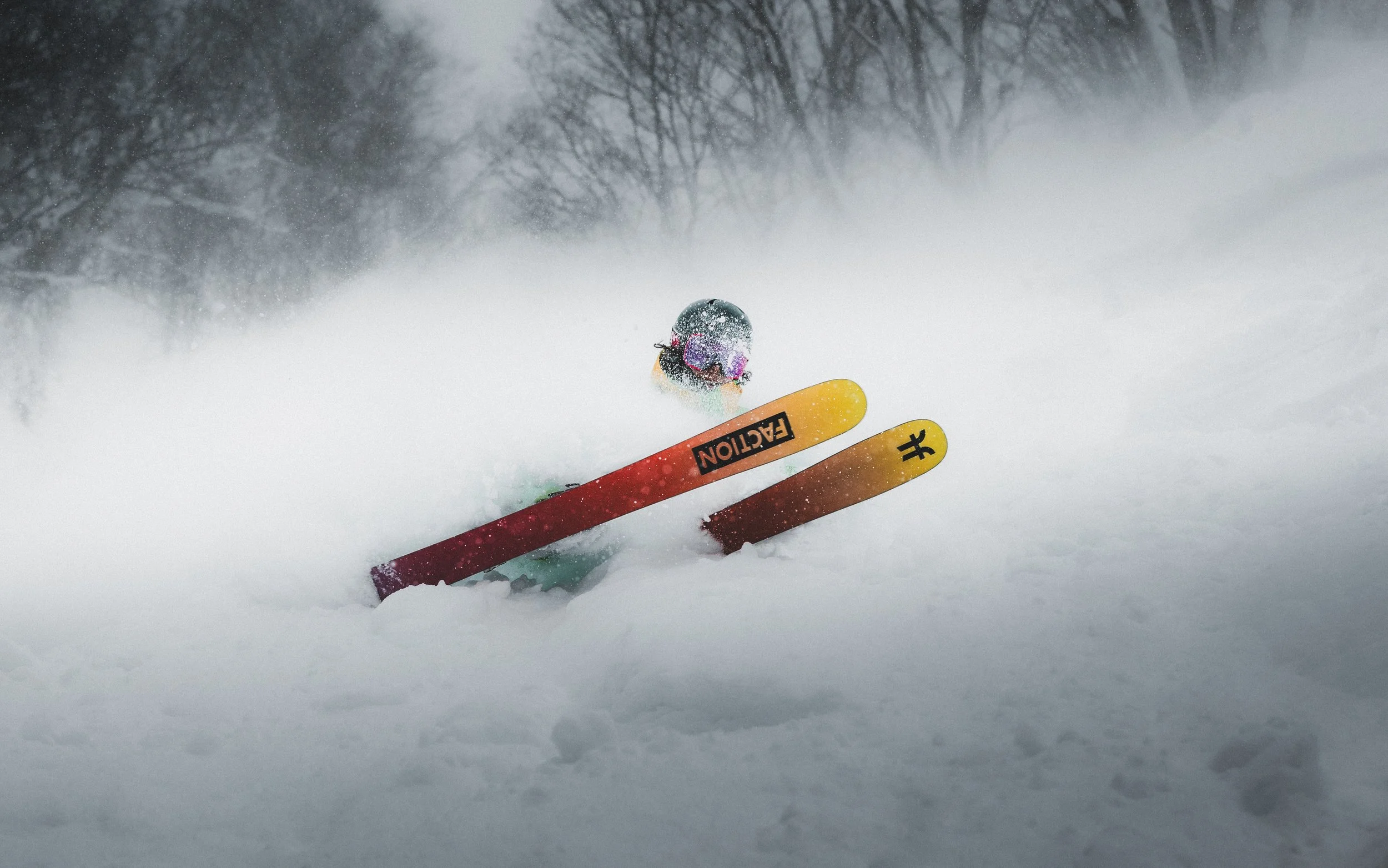 A skier with safety goggles and helmet falling in deep snow on a snowy slope with trees in the background.