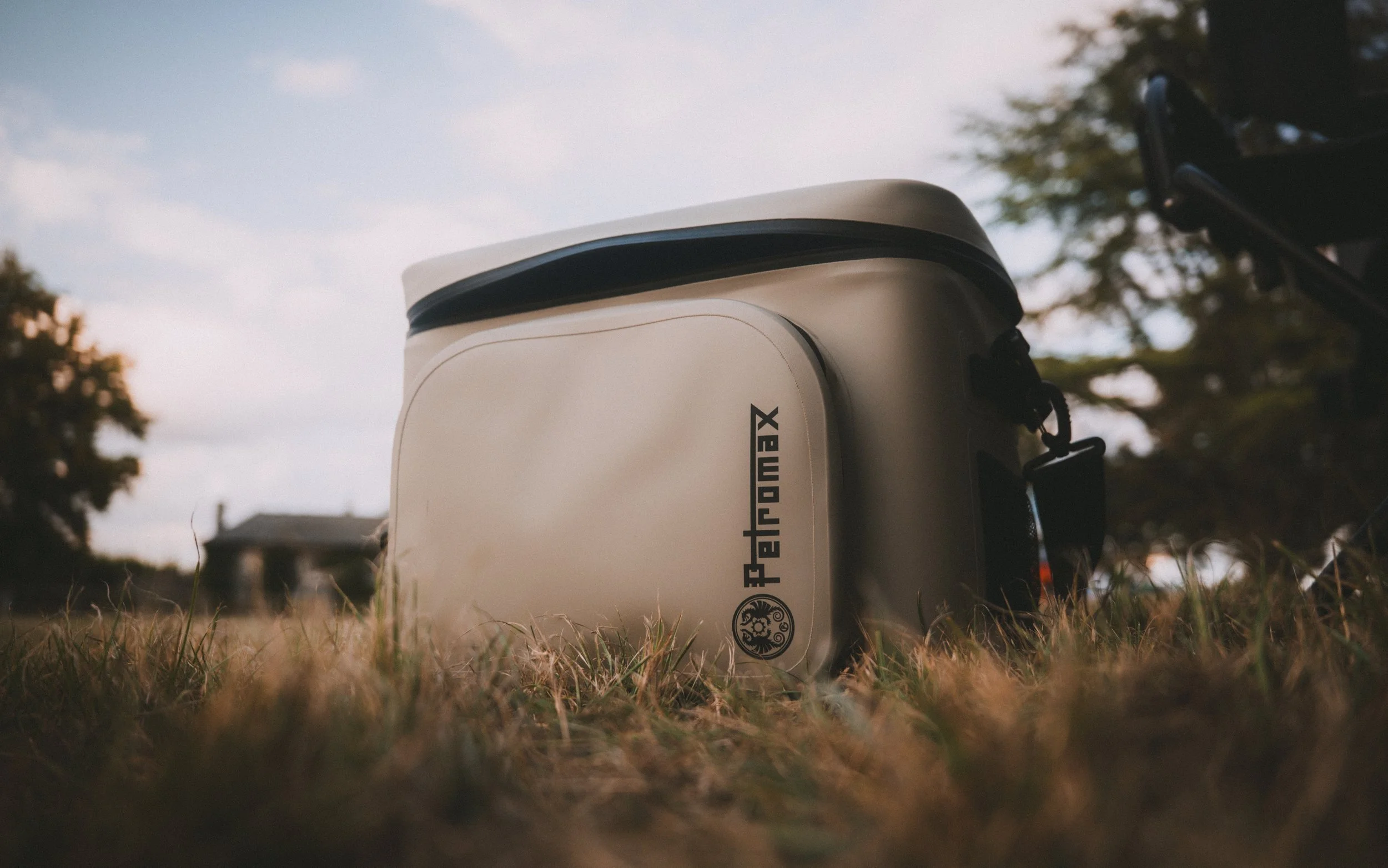 A white Petromax lantern placed on the grass outdoors with a blurred house and trees in the background.